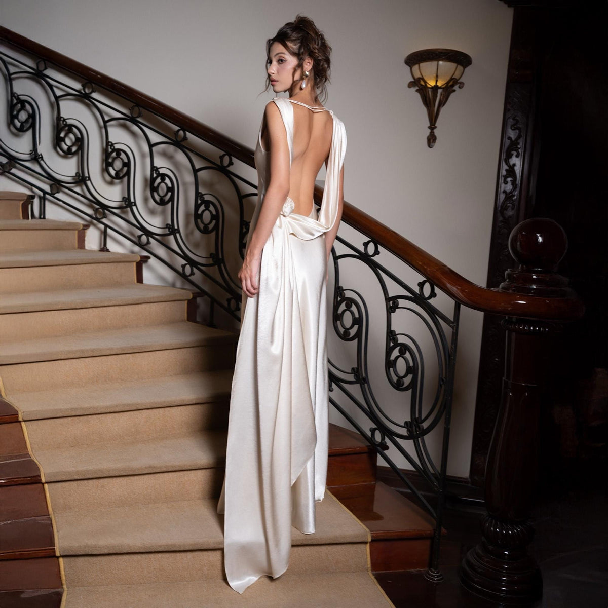 May include: A cream-colored, floor-length gown with a draped back and train. The dress is made of a silky material and is worn on a staircase with a brown handrail and black wrought iron detailing. The stairs are covered in a beige carpet.
