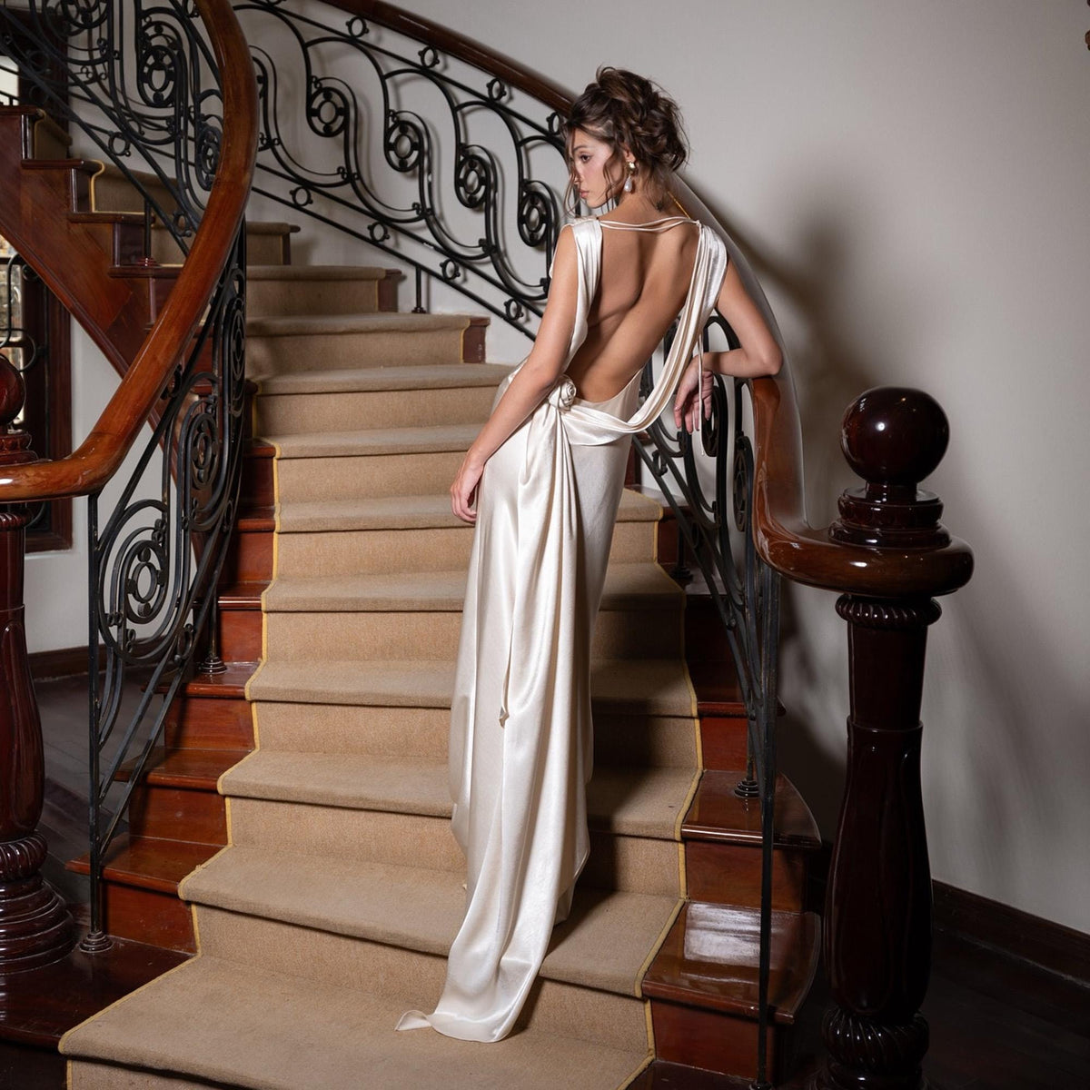 May include: A cream-colored, backless satin gown with a draped cowl neckline. The dress is floor-length and features a flowing skirt. The model is standing on a staircase with a wooden banister and wrought iron railing.