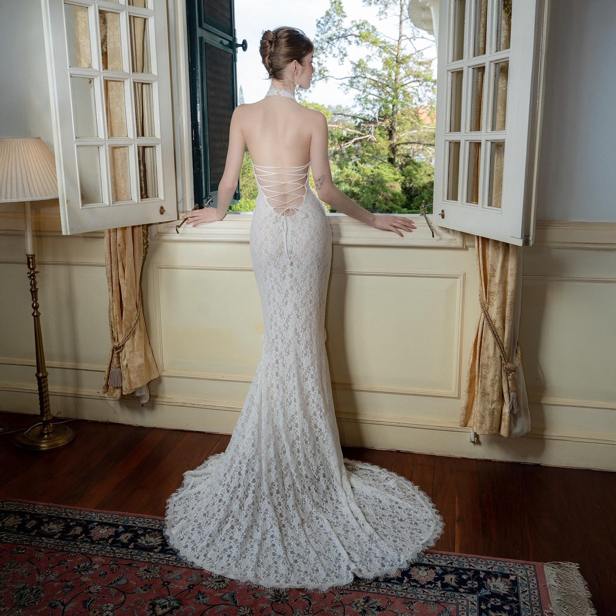 May include: A white lace wedding dress with a halter neckline and open back. The dress has a fitted silhouette that flares into a mermaid style with a long train. The bride is standing near a window.