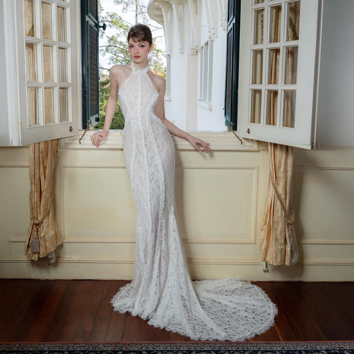 May include: A white, floor-length lace gown with a halter neckline and a mermaid silhouette. The dress features intricate floral lace detailing and a long train. The model is standing near an open window with cream-colored curtains.