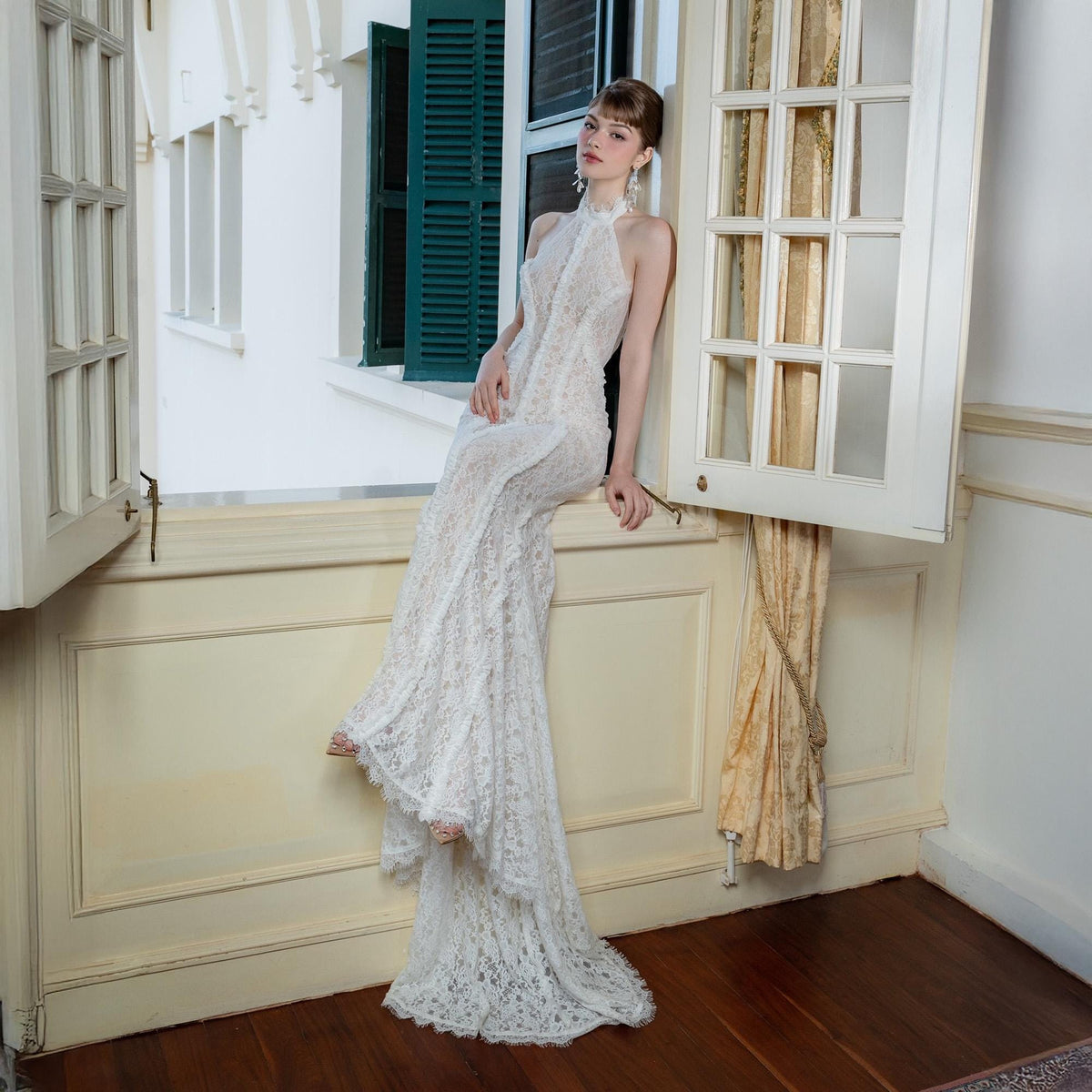 May include: A white lace halter neck gown with a flowing train. The dress features a high neckline and intricate lace detailing. The model is seated near a window with green shutters and a cream-colored wall.