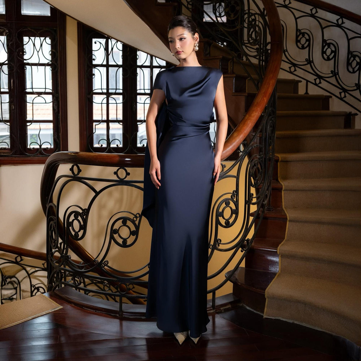 May include: A navy blue, floor-length gown with a draped neckline and short sleeves. The dress features a flowing silhouette and is made of a shiny, silky material. The woman is standing on a wooden staircase.