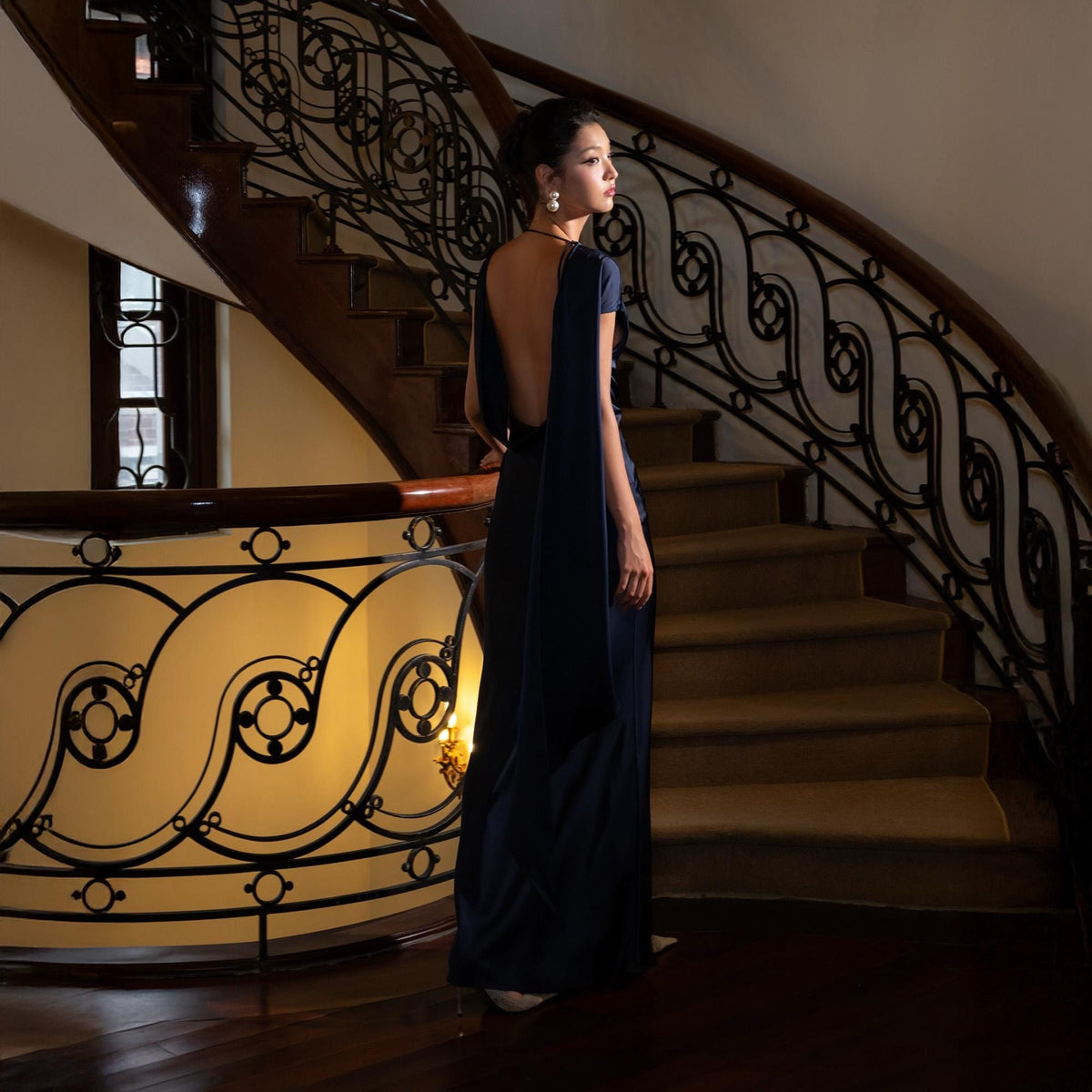May include: A woman in a navy blue, backless evening gown stands on a wooden staircase with ornate wrought iron railings. The dress has a flowing cape detail and a simple, elegant design. The setting is a well-lit, classic interior.