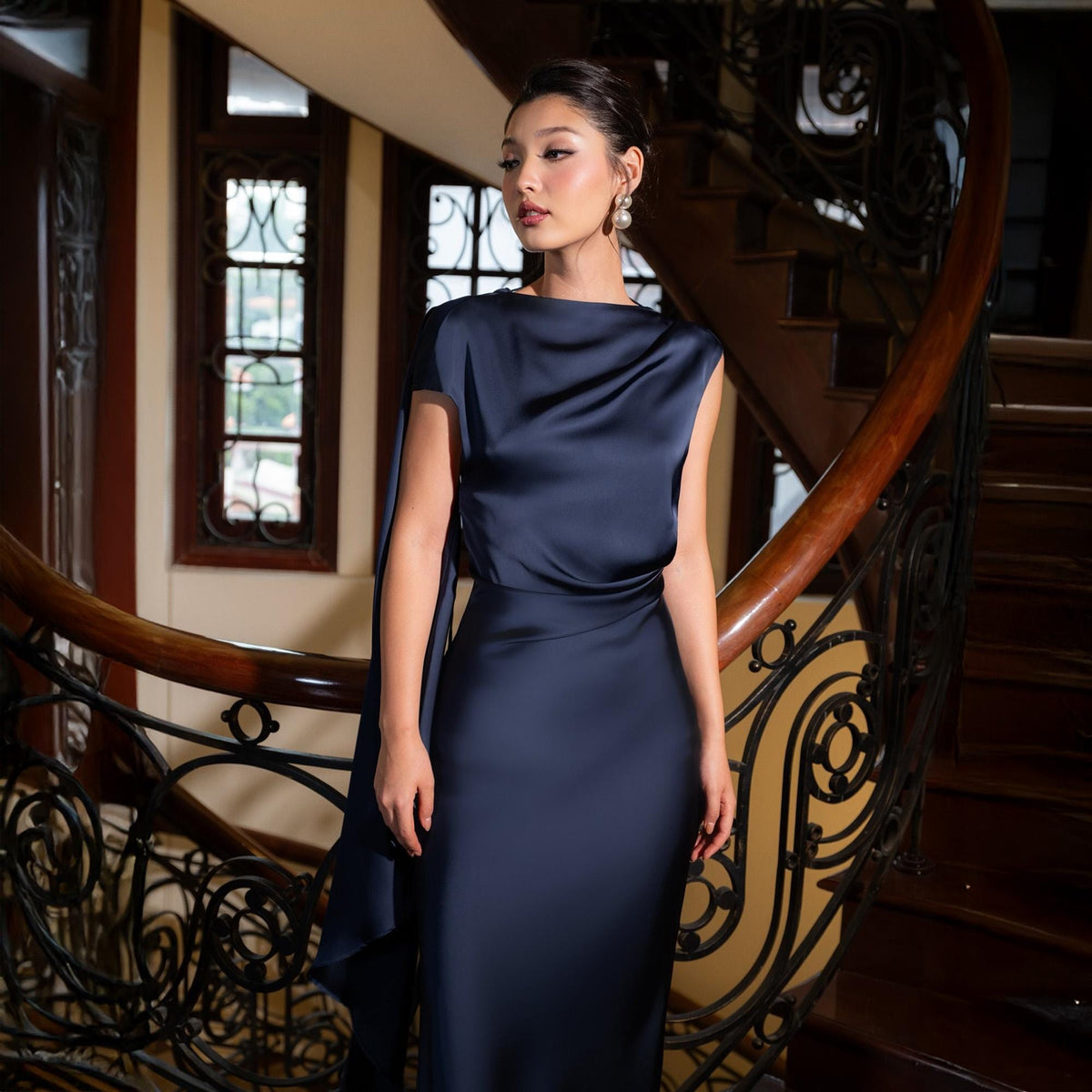 May include: A navy blue satin gown with a draped neckline and a flowing cape detail. The dress is floor-length and form-fitting, creating an elegant silhouette. The model is wearing pearl earrings. The background features a wooden staircase.