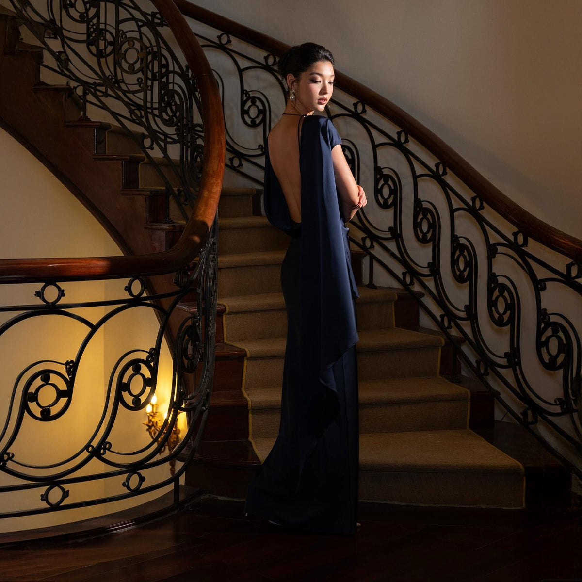 May include: A woman in a navy blue, open-back evening gown stands on a wooden staircase with ornate wrought iron railings. The dress features a flowing cape-like design and a floor-length skirt. The setting is a well-lit interior.