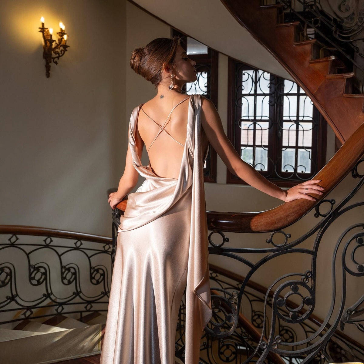 May include: A champagne-colored satin gown with a low back and draped detailing. The dress features delicate straps and a flowing skirt. The woman is standing on a wooden staircase with ornate metal railings.