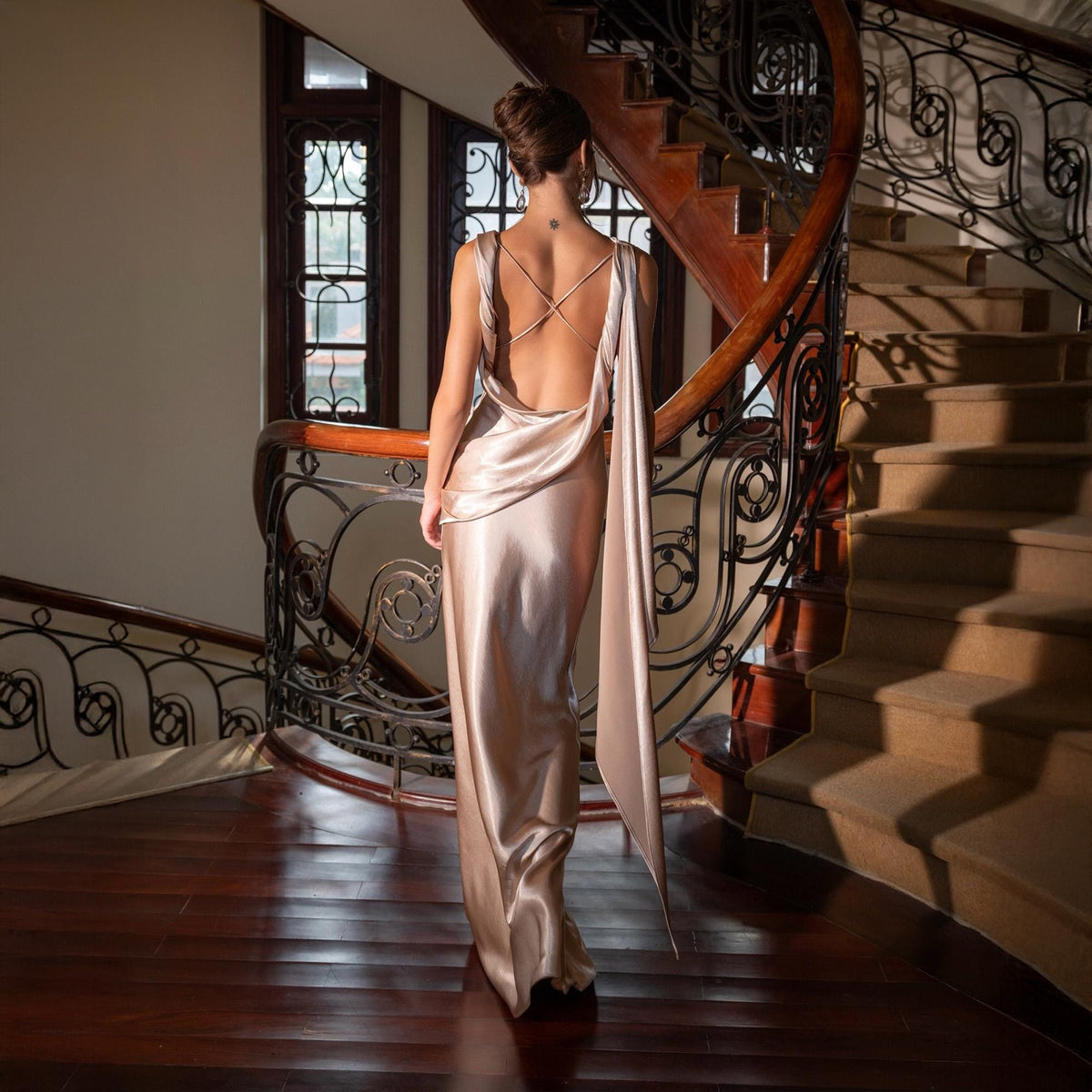 May include: A long, champagne-colored satin gown with a low back and draped fabric detail. The dress is worn in a grand, well-lit space with a winding staircase and dark wood floors. The gown's design features delicate straps across the back.