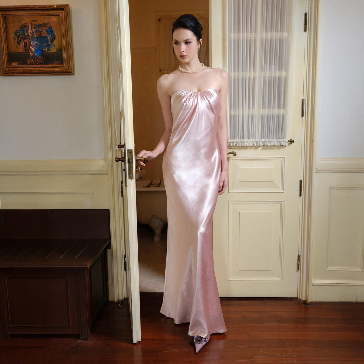 May include: A strapless, floor-length, pale pink satin gown. The dress features a gathered bodice and a flowing skirt. The model is wearing a pearl necklace. The dress is elegant and suitable for formal events.