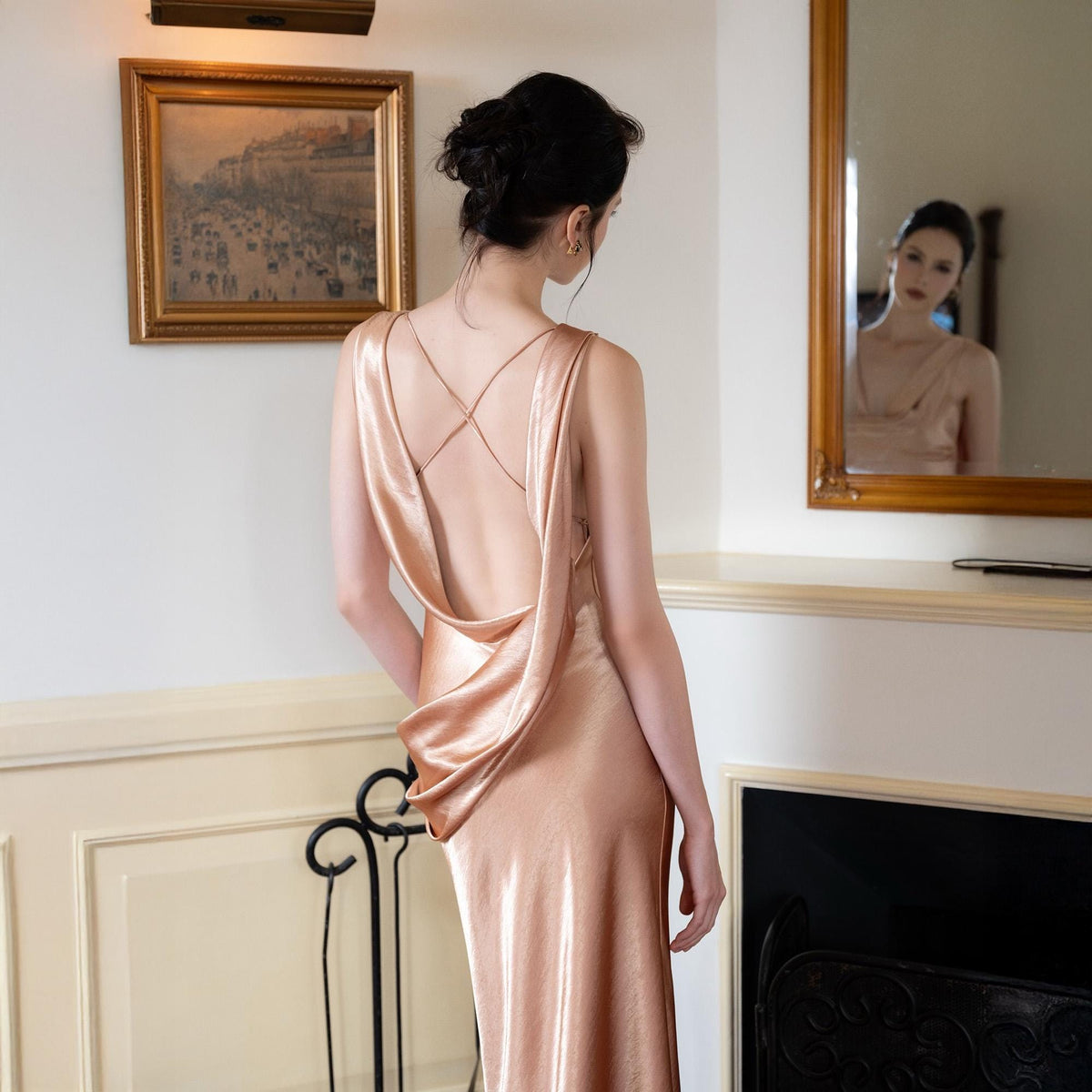 May include: A peach-colored satin dress with a cowl neckline and a low back. The dress has delicate spaghetti straps that cross in the back. The model is standing in front of a mirror and a fireplace.