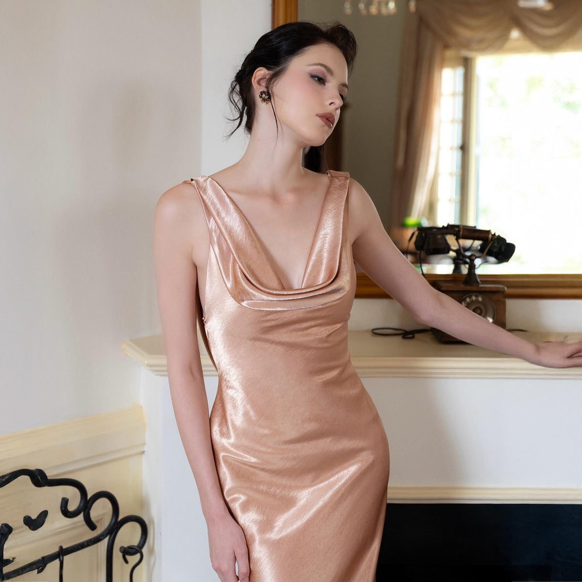 May include: A blush-colored satin slip dress with a cowl neckline. The dress has a sleeveless design and a flowing silhouette. The model is wearing the dress in a room with a fireplace and a mirror.