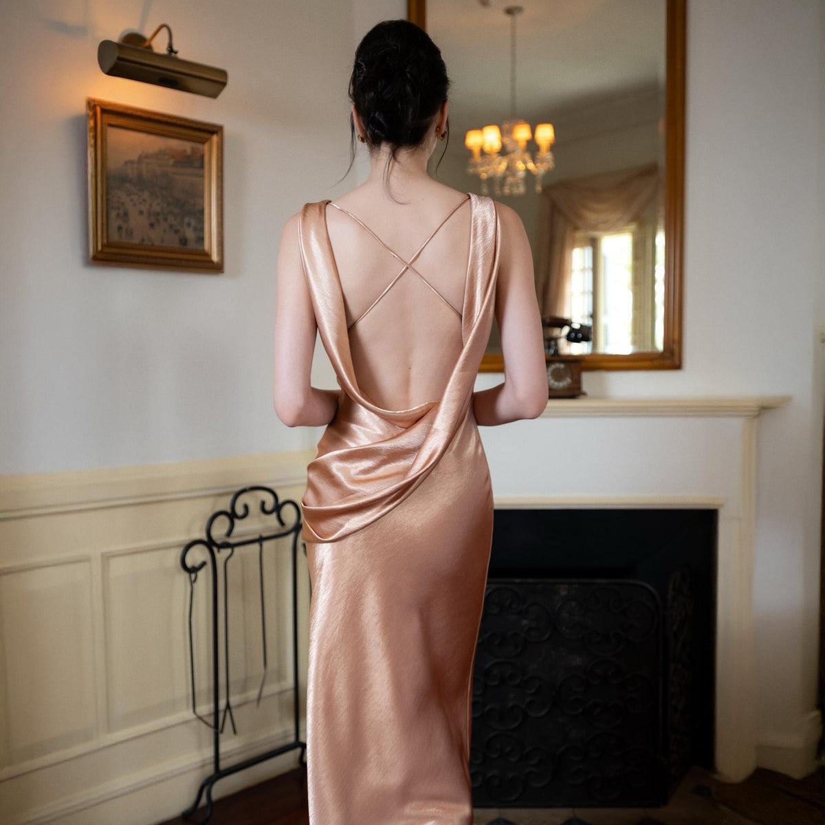 May include: A rose gold satin dress with a cowl neckline and a low back. The straps cross in the back. The dress is floor-length and has a slight sheen. The dress is elegant and suitable for formal events.