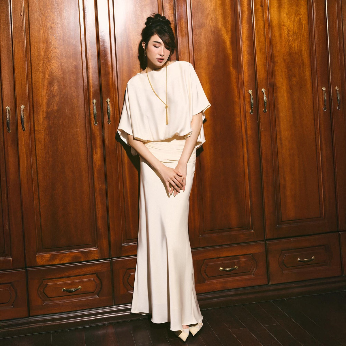 May include: A cream-colored, two-piece formal outfit. The top is a short-sleeved cape-style blouse, and the bottom is a floor-length, form-fitting skirt. A gold necklace with a long pendant is worn. The background is a dark wood wardrobe.