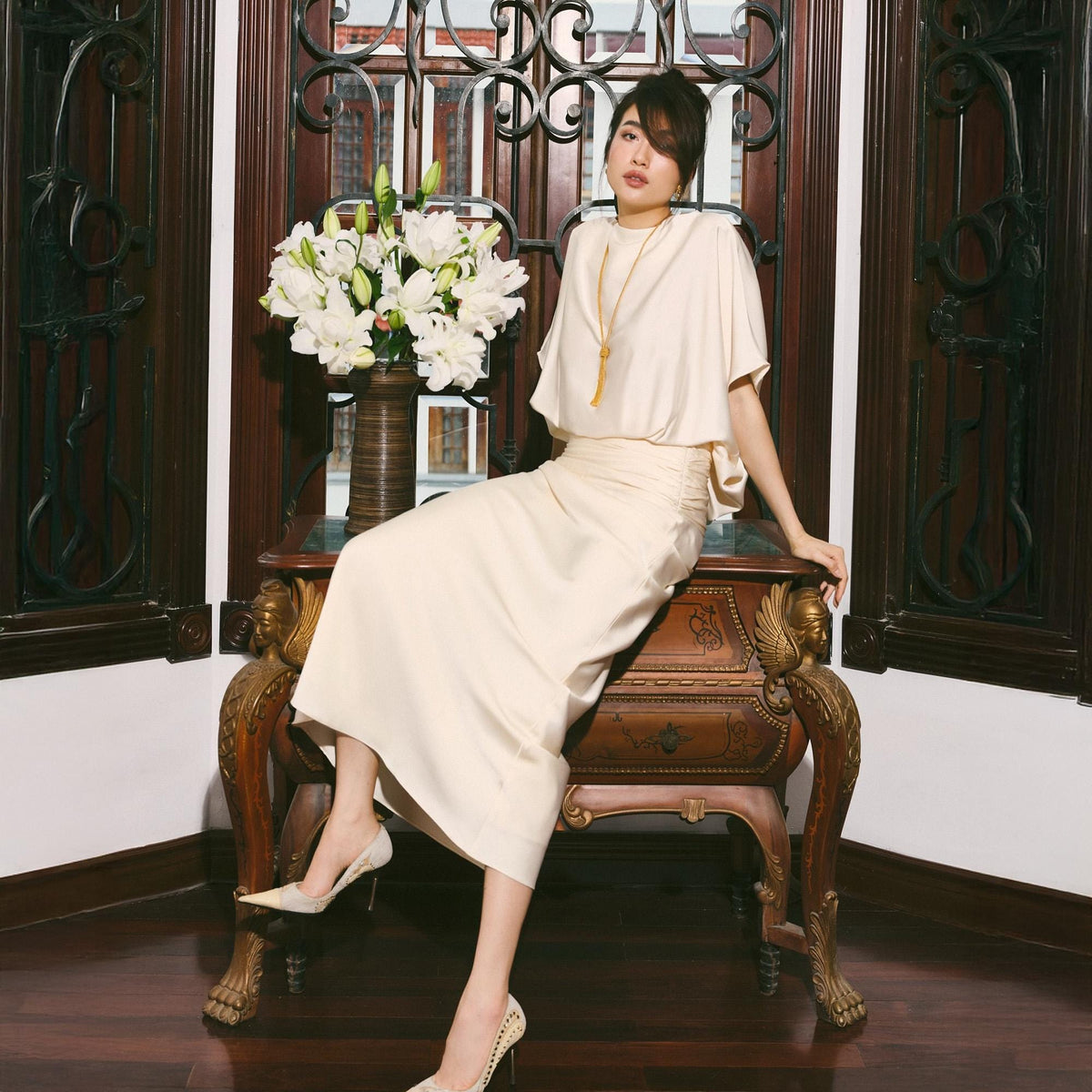 May include: A woman wearing a cream-colored outfit, including a top, skirt, and heels, sits on a vintage wooden table. The table has ornate carvings and is positioned near a window with decorative ironwork. A vase of white flowers is also visible.