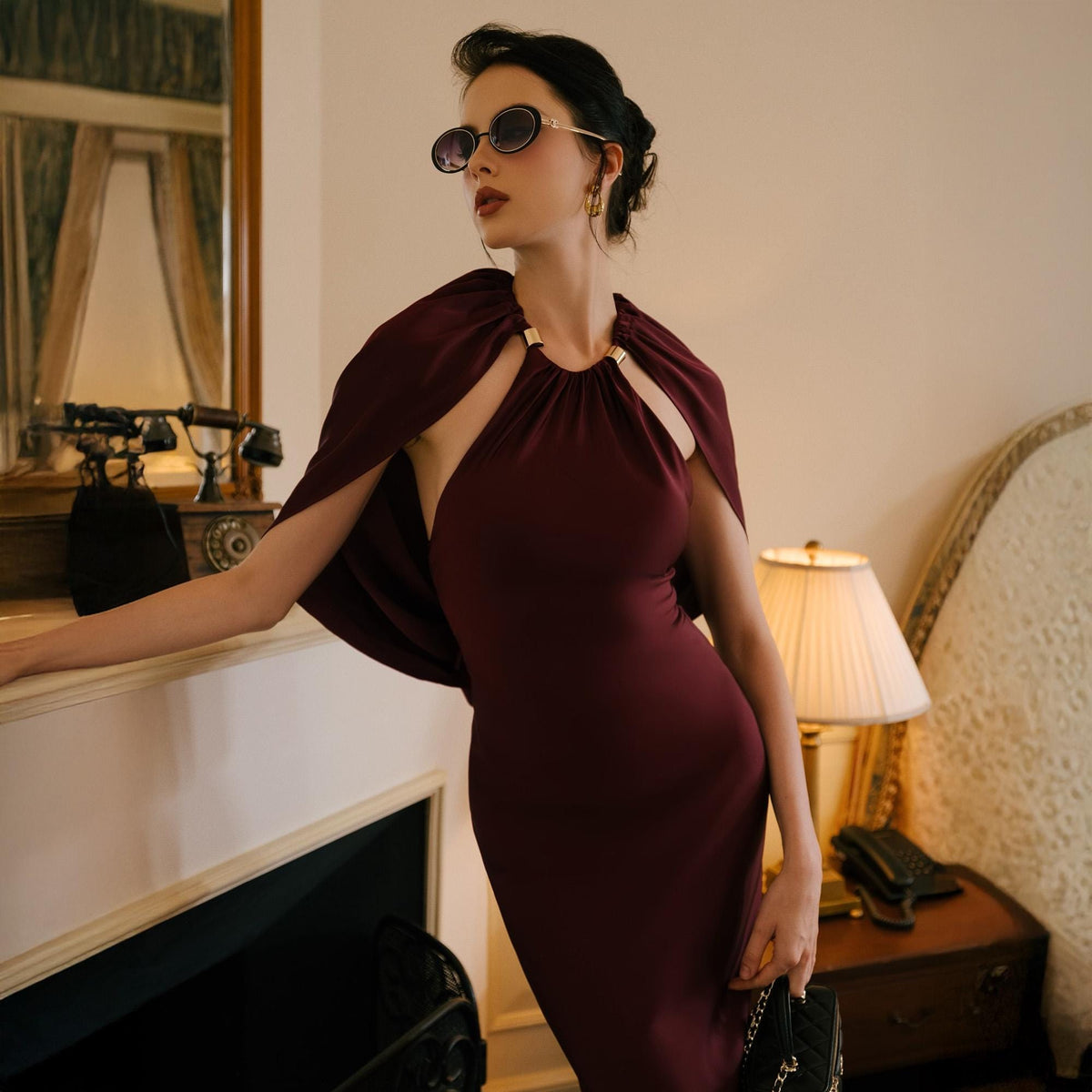 May include: A burgundy halter dress with a cape detail. The dress features a keyhole neckline with gold accents. The model is wearing sunglasses and gold earrings. The dress is form-fitting and elegant, suitable for formal events.