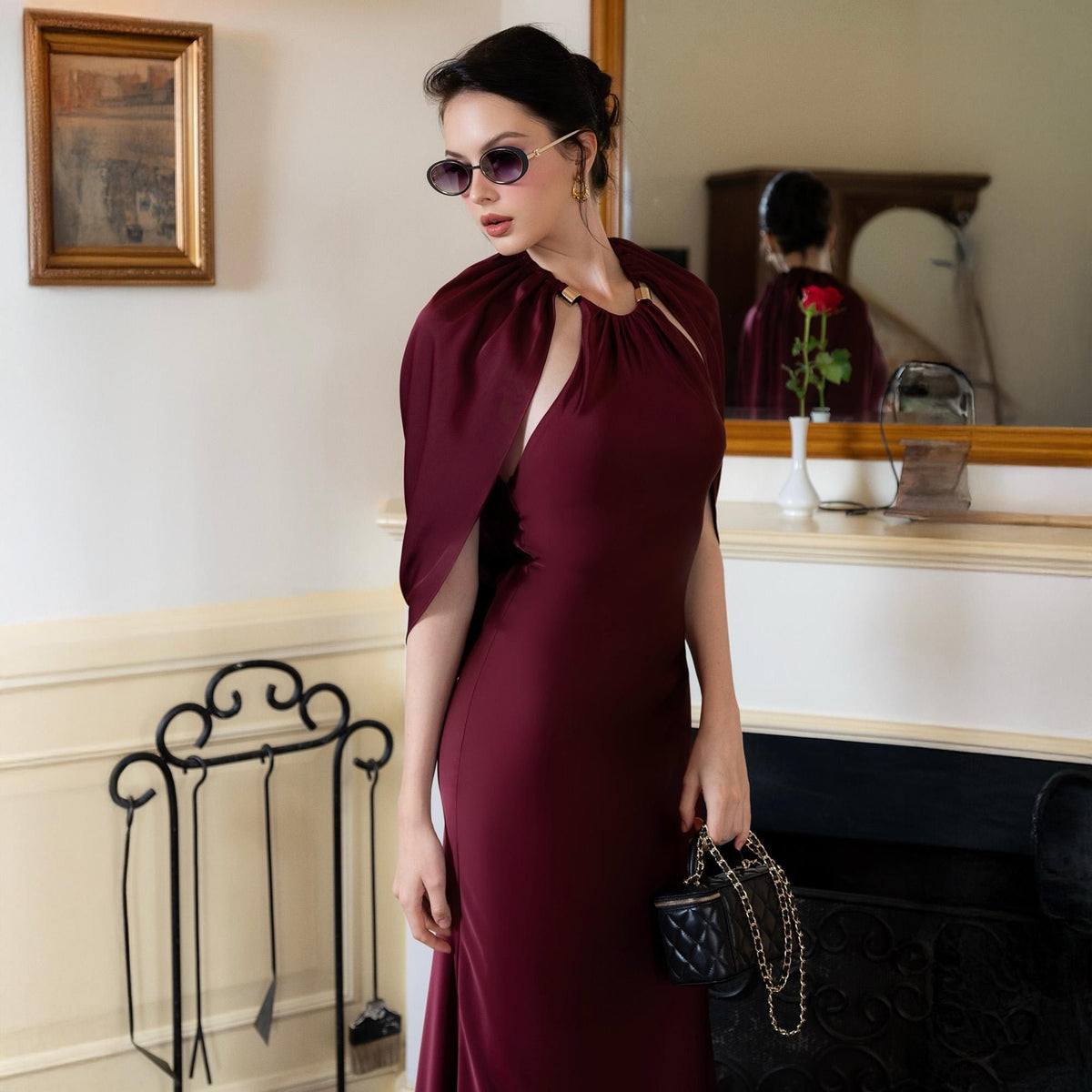 May include: A burgundy formal gown with a matching cape. The dress has a fitted silhouette and a high neckline. The cape is attached at the shoulders and drapes over the arms. A small black quilted handbag with a gold chain is held in the hand.