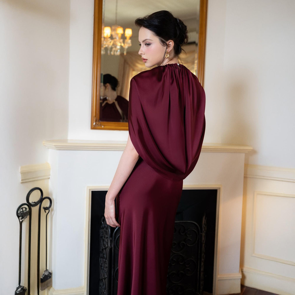 May include: A burgundy satin gown with a draped cape-like top and a floor-length skirt. The dress is worn by a person standing near a fireplace with a mirror reflecting the person's image. The room has white walls and a gold-framed mirror.