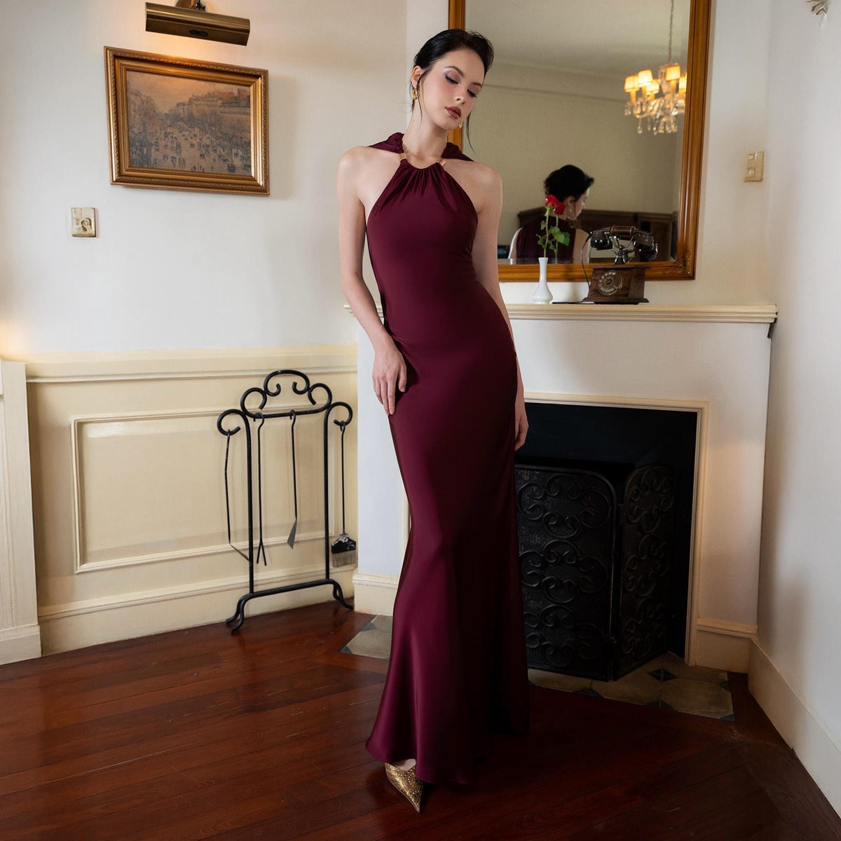 May include: A burgundy halter neck gown with a floor-length silhouette. The dress features a fitted bodice and a flared skirt. The model is wearing gold-colored shoes. The background includes a fireplace, mirror, and a decorative metal stand.
