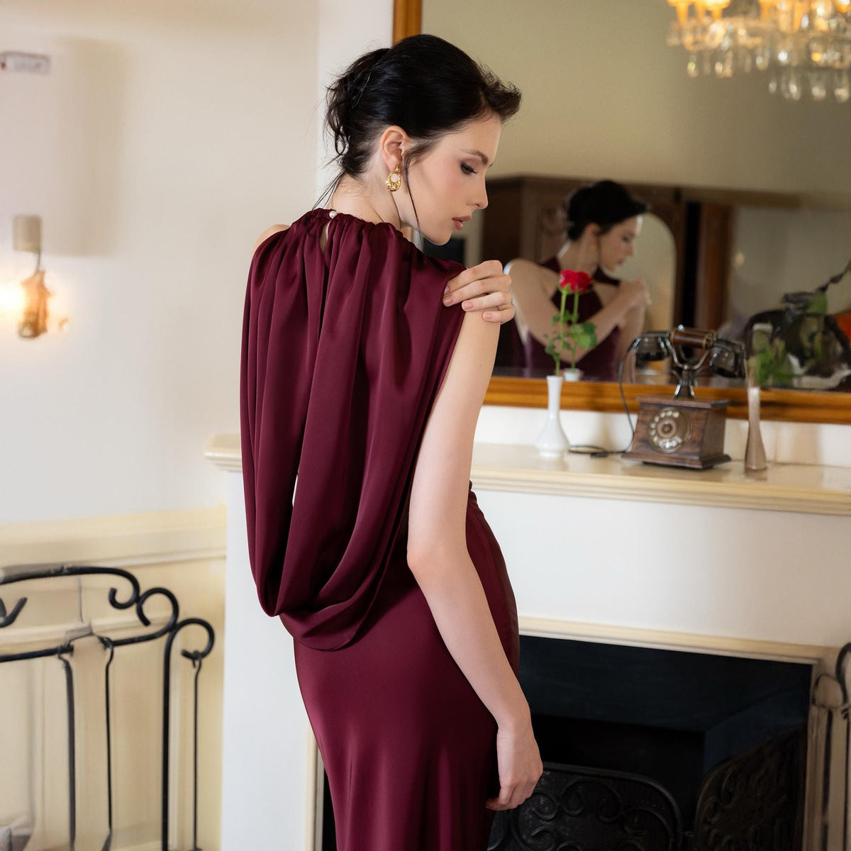 May include: A burgundy-colored, floor-length dress with a draped cowl neckline. The dress has a sleek, form-fitting silhouette. The model is wearing gold earrings. The background includes a fireplace, mirror, and a vintage telephone.