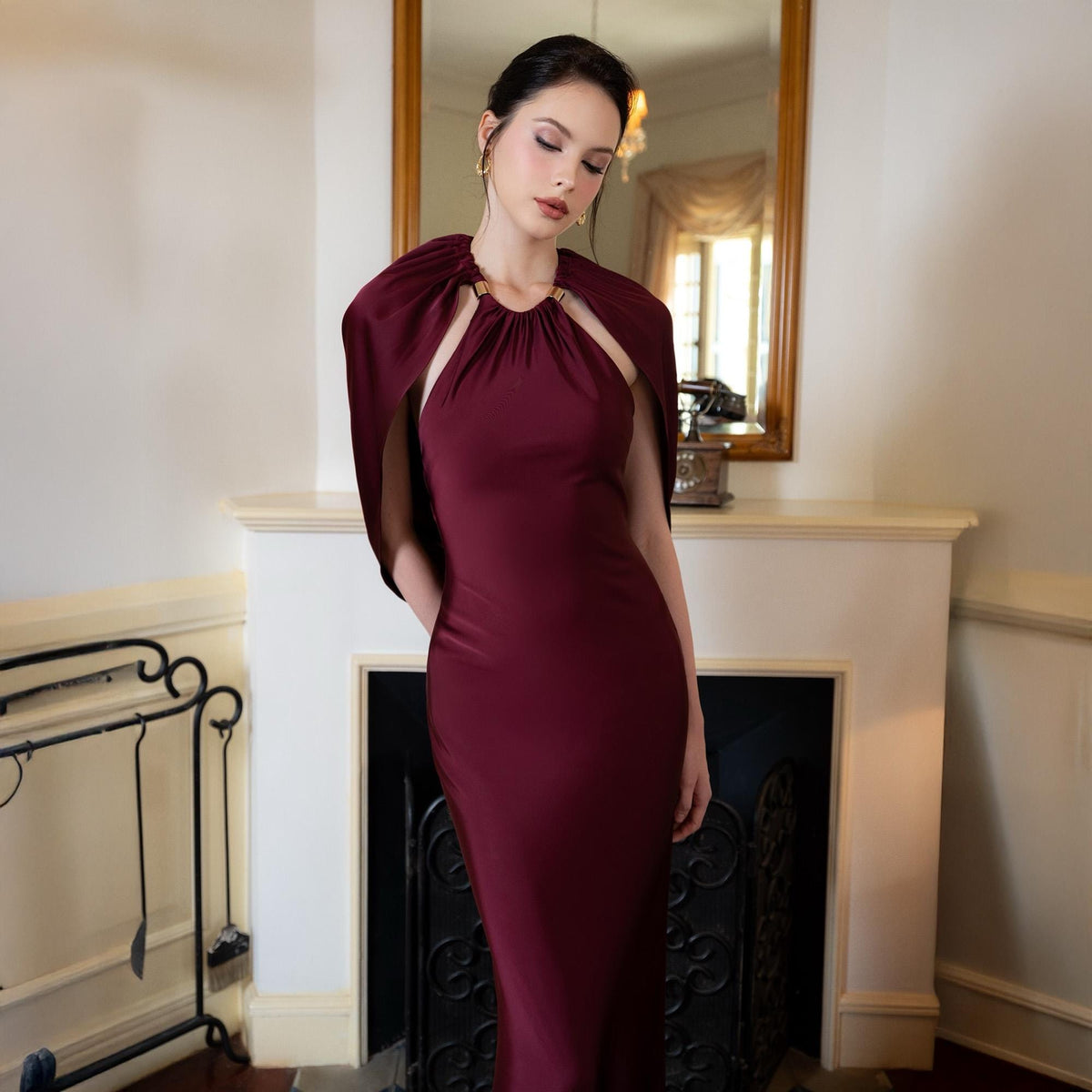 May include: A burgundy-colored formal gown with a cape-like overlay. The dress features a halter neckline with a gold-colored accent. The cape drapes over the shoulders, creating a sophisticated silhouette. The dress is floor-length and form-fitting.