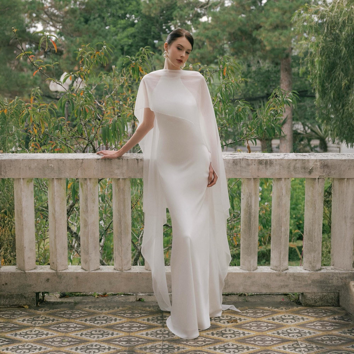 May include: A white bridal gown with a high neckline and a flowing, floor-length skirt. The dress is paired with a sheer, cape-like overlay. The model is standing on a balcony with a stone railing, with greenery in the background.
