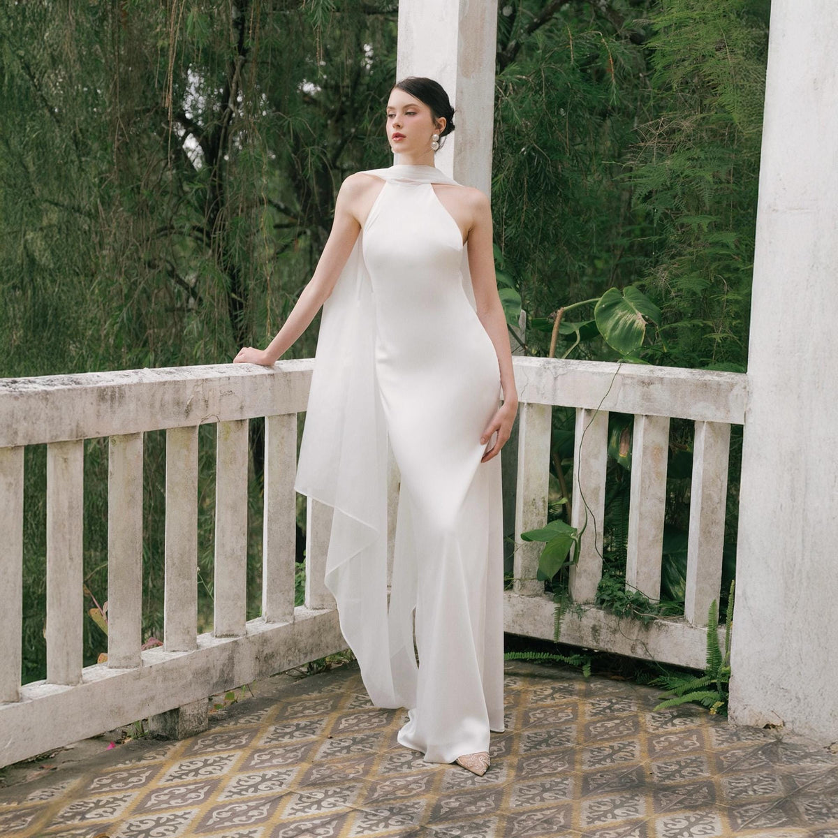 May include: A white halter-neck jumpsuit with a flowing, sheer cape. The elegant bridal outfit features a fitted bodice and wide-leg pants. The model is standing on a balcony with a white railing, surrounded by greenery.