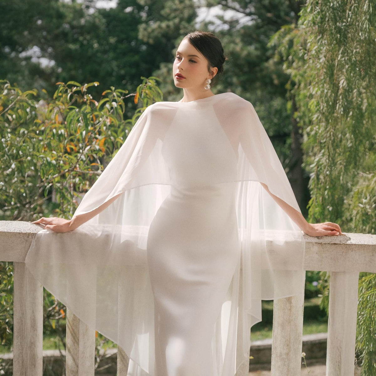 May include: A white wedding dress with a sheer cape. The dress is a simple, elegant design with a fitted bodice and a flowing skirt. The cape drapes over the shoulders and arms, adding a touch of sophistication. The bride is standing on a white balcony with green foliage in the background.