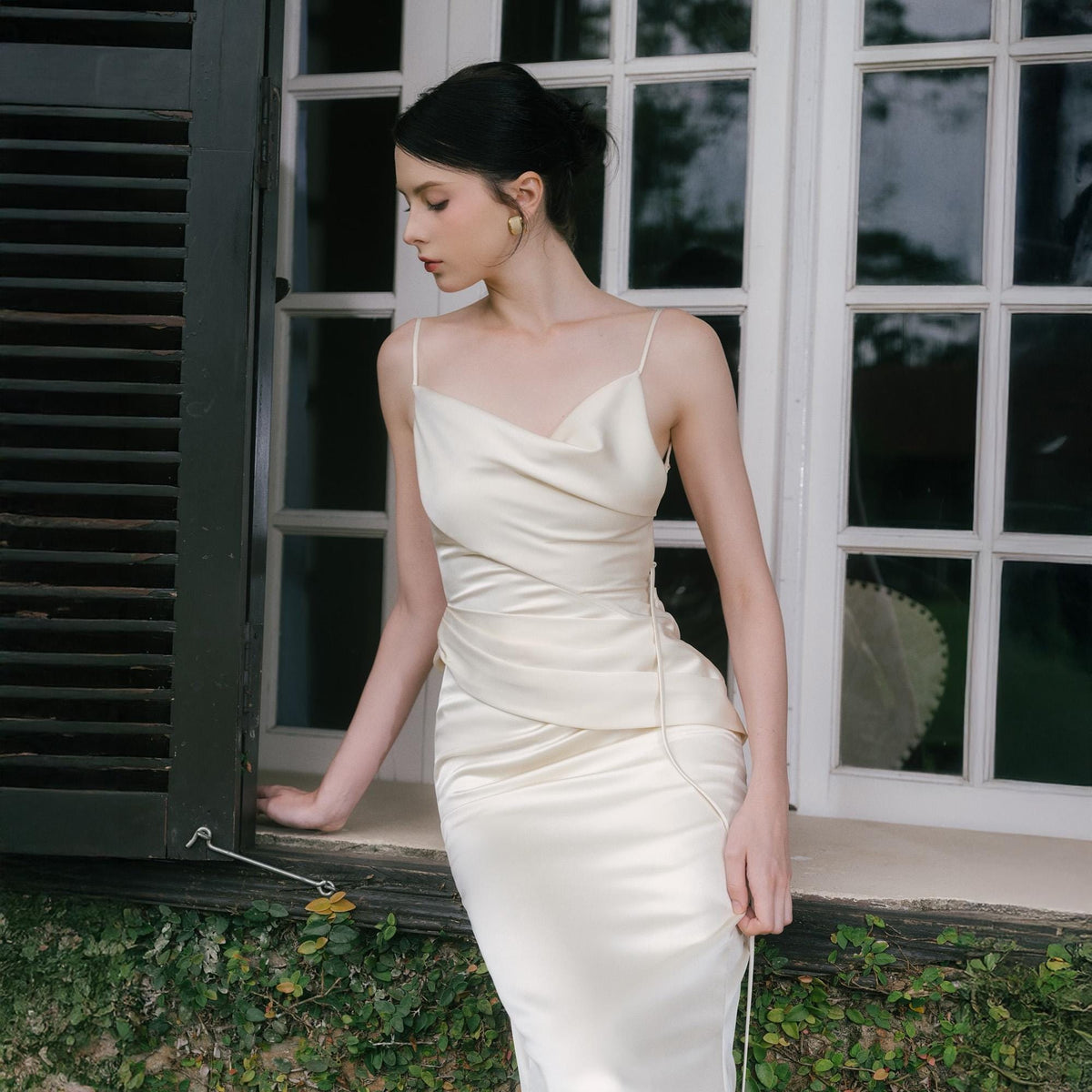 May include: Ivory satin slip dress with a cowl neckline and spaghetti straps. The dress features a draped bodice and a fitted silhouette. The model is standing near a window with white frames and green shutters.