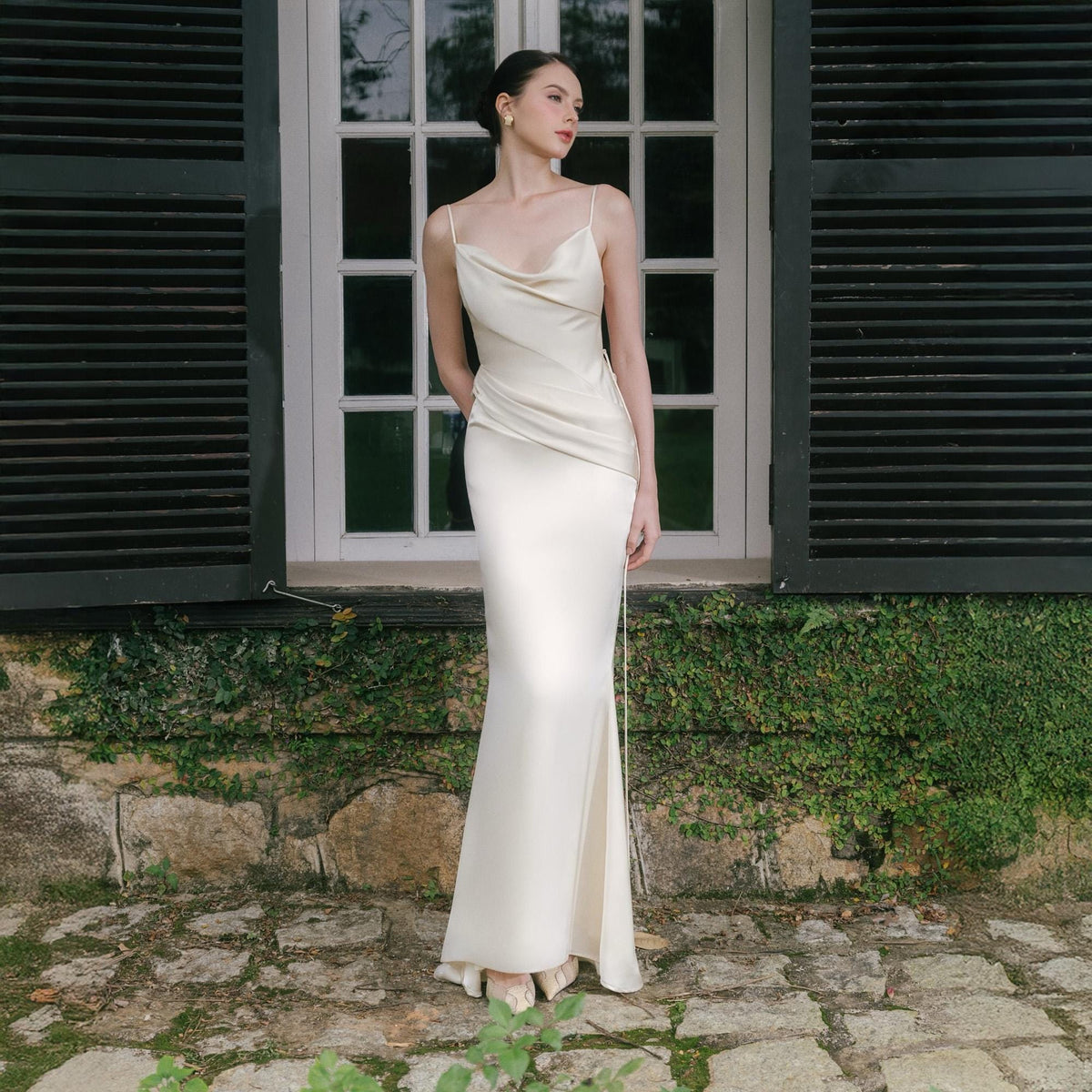 May include: A floor-length ivory satin gown with a cowl neckline and spaghetti straps. The dress features a fitted silhouette that flares out slightly at the hem. The model is standing in front of a window with dark shutters.