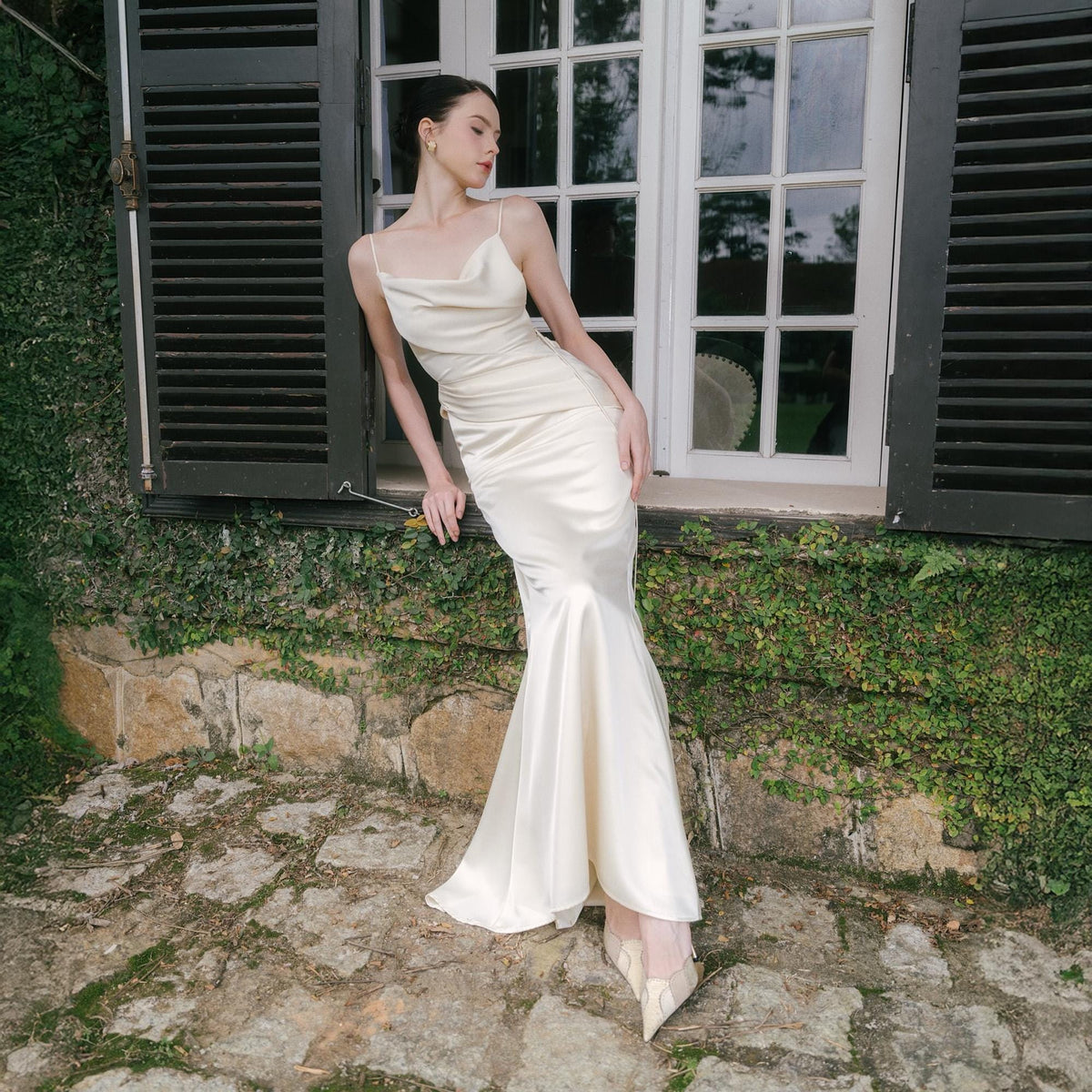 May include: A cream-colored, floor-length gown with a cowl neckline and spaghetti straps. The dress has a fitted bodice and flares out at the hem. The model is leaning against a window with black shutters, surrounded by greenery.