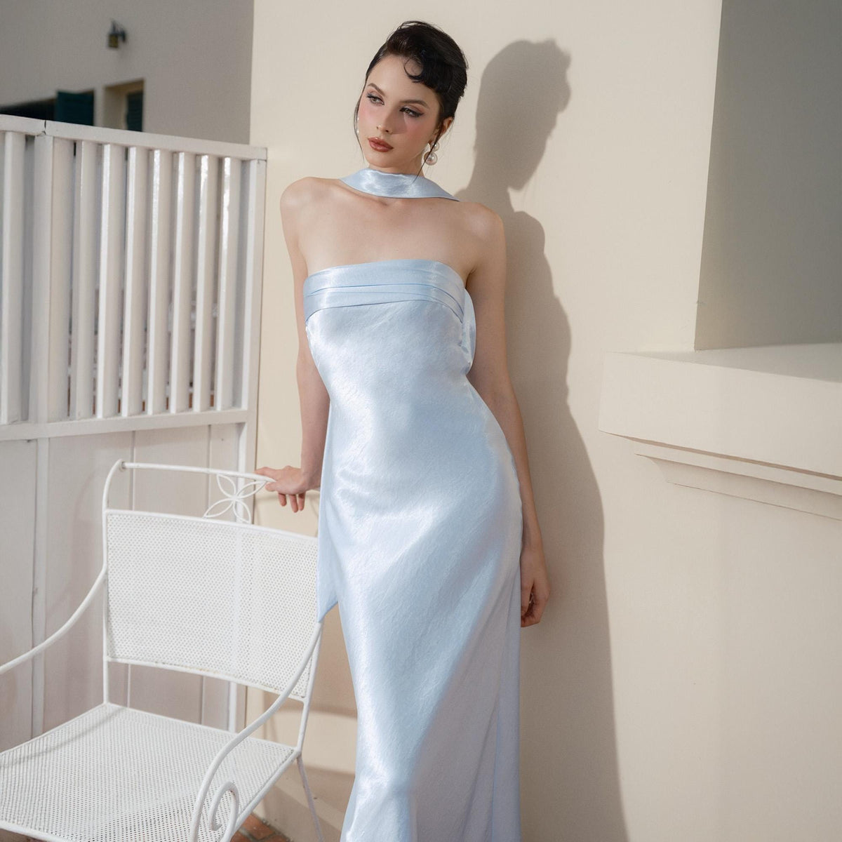 May include: A light blue strapless dress with a matching neck scarf. The dress has a side slit and a fitted silhouette. A white metal chair is in the foreground.