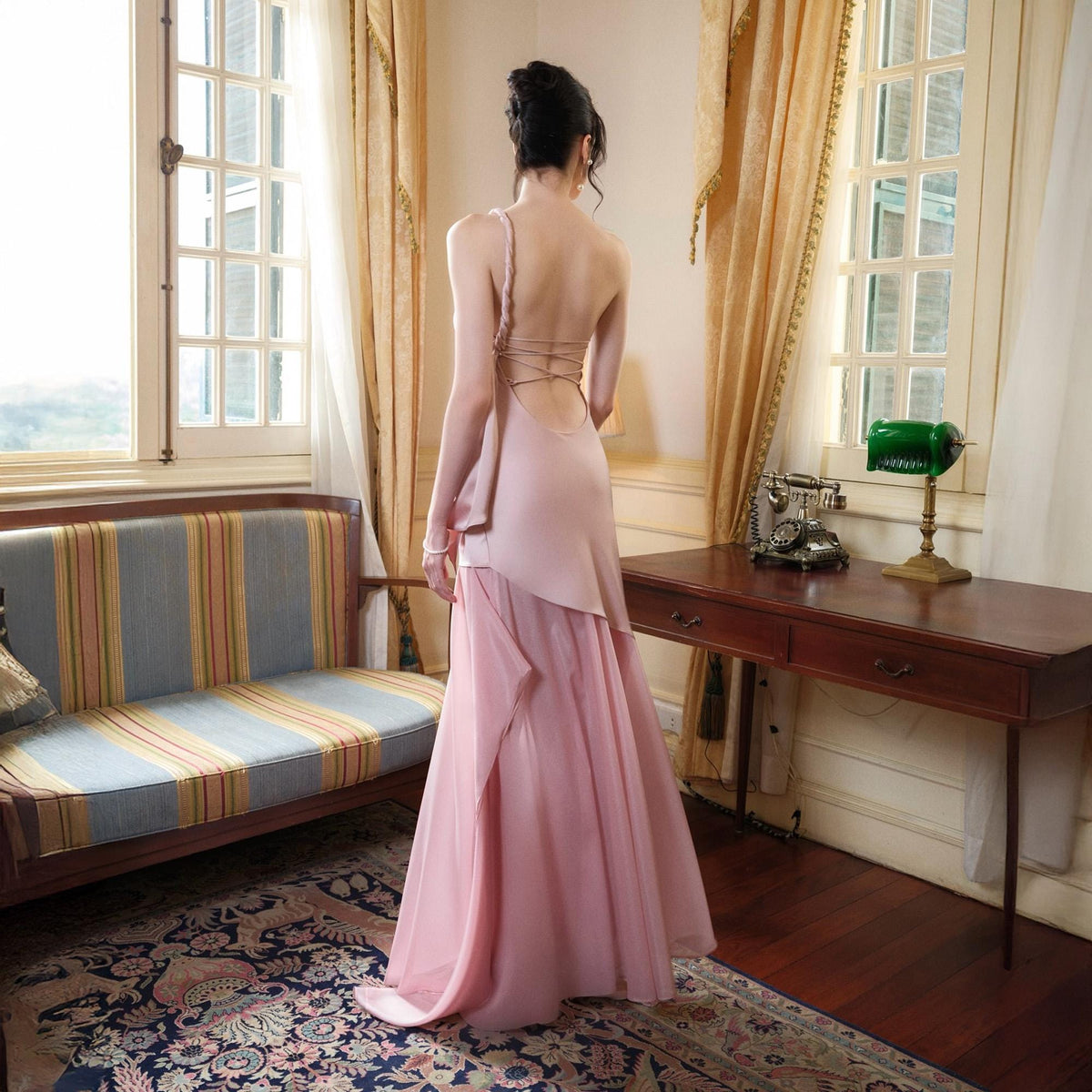 May include: A light pink formal gown with a flowing skirt and a draped bodice. The back of the dress features delicate strap detailing. The dress is worn in a room with a vintage sofa and a wooden desk.
