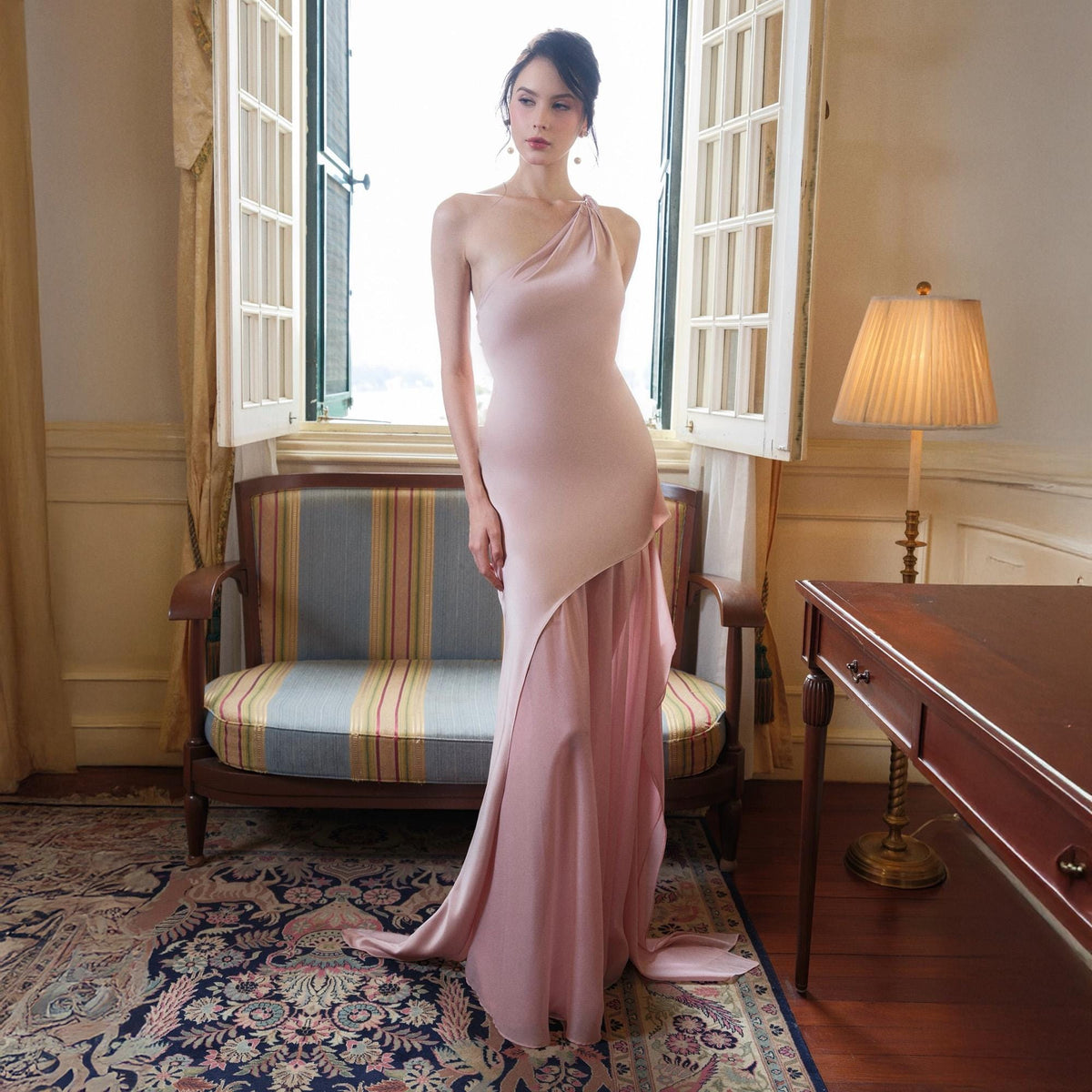 May include: A blush-colored, one-shoulder formal gown with a flowing skirt. The dress features a fitted bodice and a draped, asymmetrical skirt. The model is standing in front of a window, with a vintage sofa and rug in the background.