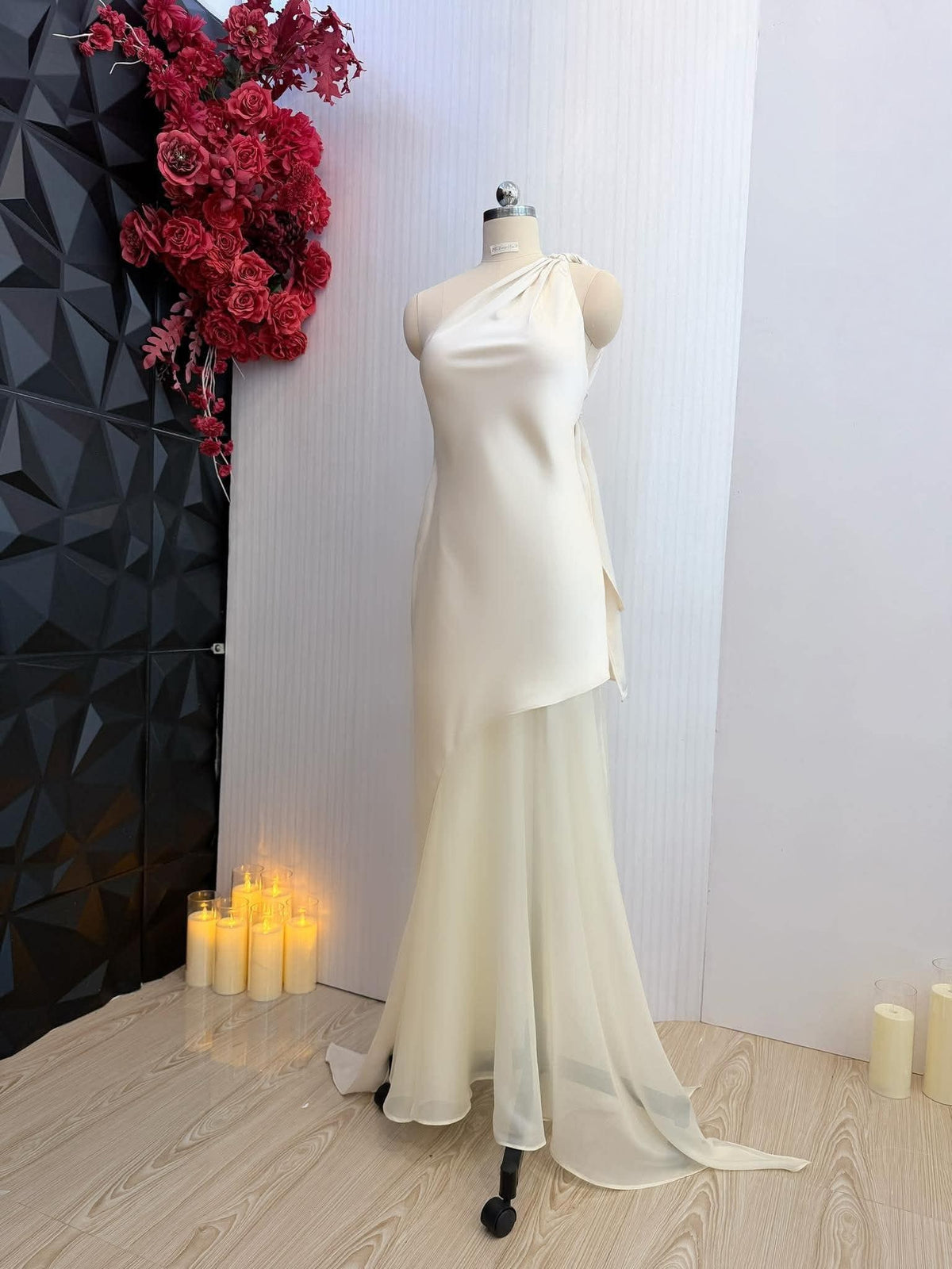 May include: Ivory one-shoulder wedding dress on a mannequin. The dress features a draped bodice and a flowing, sheer skirt. The dress is displayed in a studio setting with red flowers and candles.