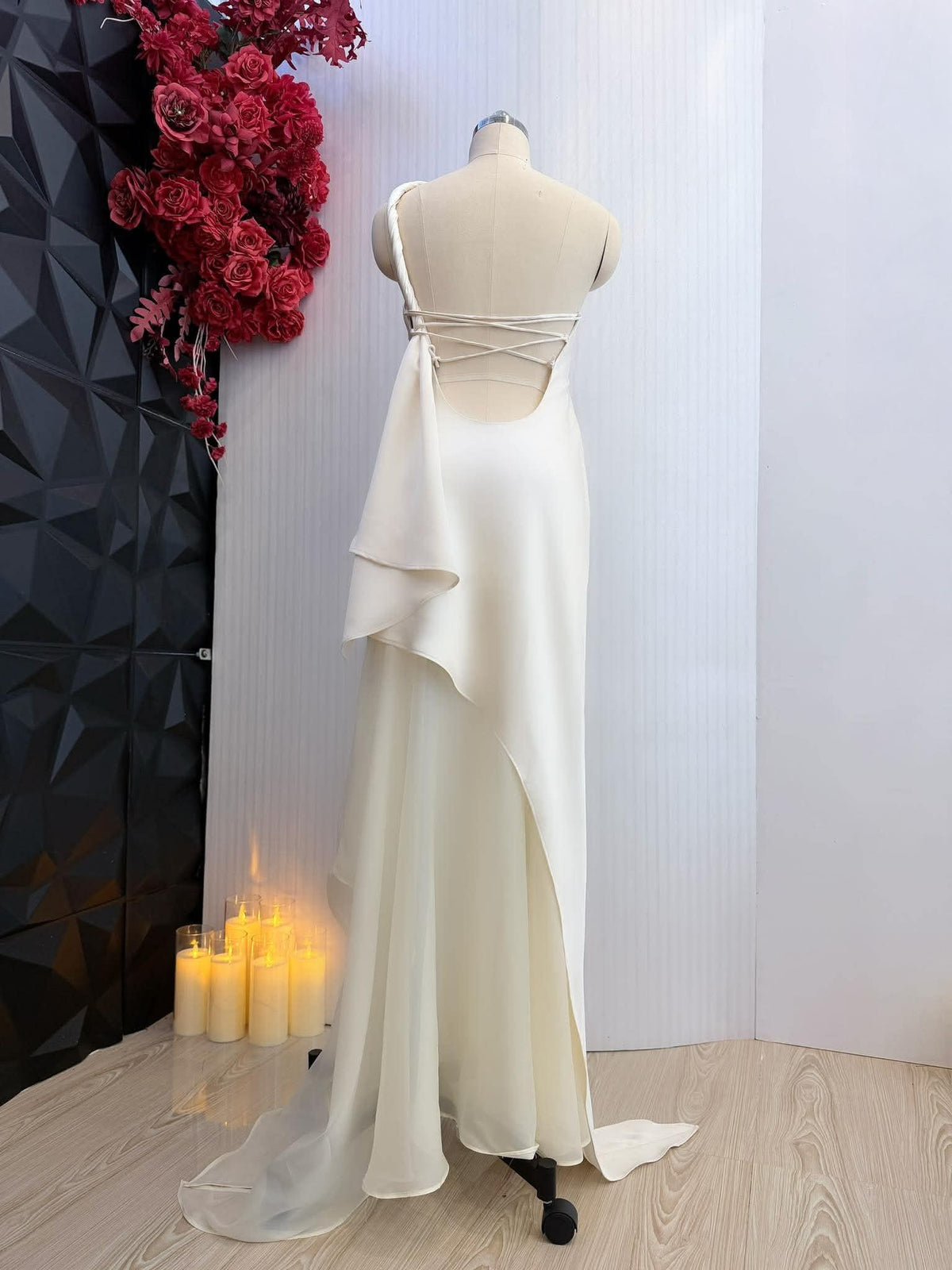 May include: Ivory-colored formal gown on a mannequin. The dress features a one-shoulder design with a crisscross back and a flowing skirt with a high slit. A sheer, layered fabric drapes from the shoulder. The dress has a train.