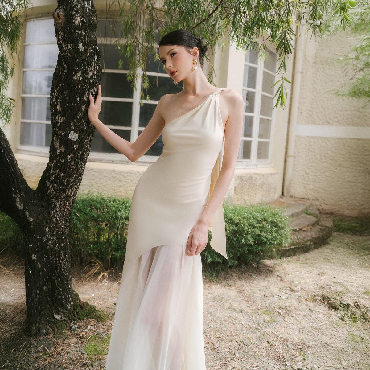 May include: A cream-colored, one-shoulder dress with a flowing skirt. The dress features a draped design and a sheer, layered skirt. The model is standing near a tree in an outdoor setting. The dress is elegant and suitable for formal events.