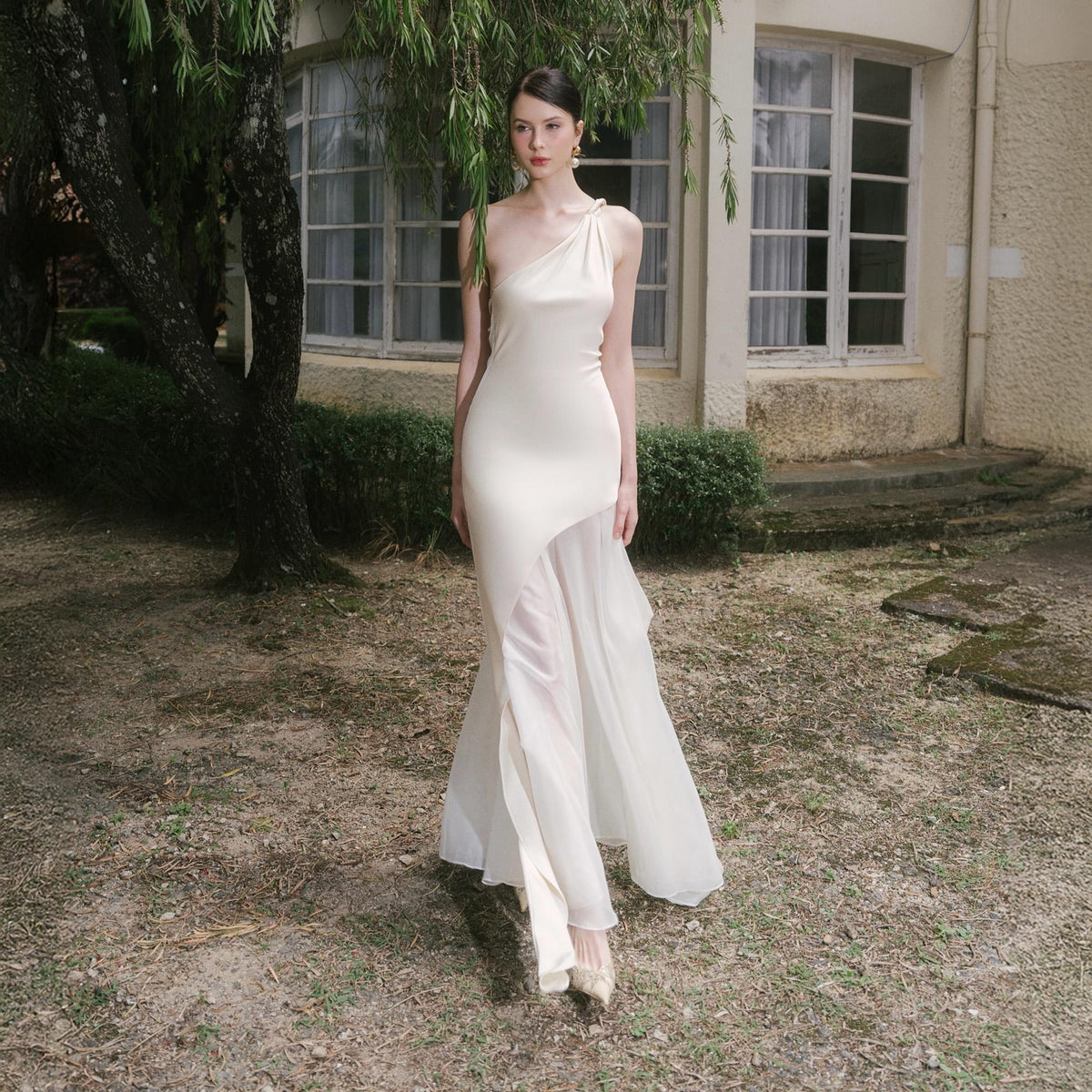 May include: A cream-colored, one-shoulder formal gown with a flowing chiffon skirt. The dress features a fitted bodice and a draped neckline. The model is standing outdoors in a garden setting, wearing matching cream-colored heels.