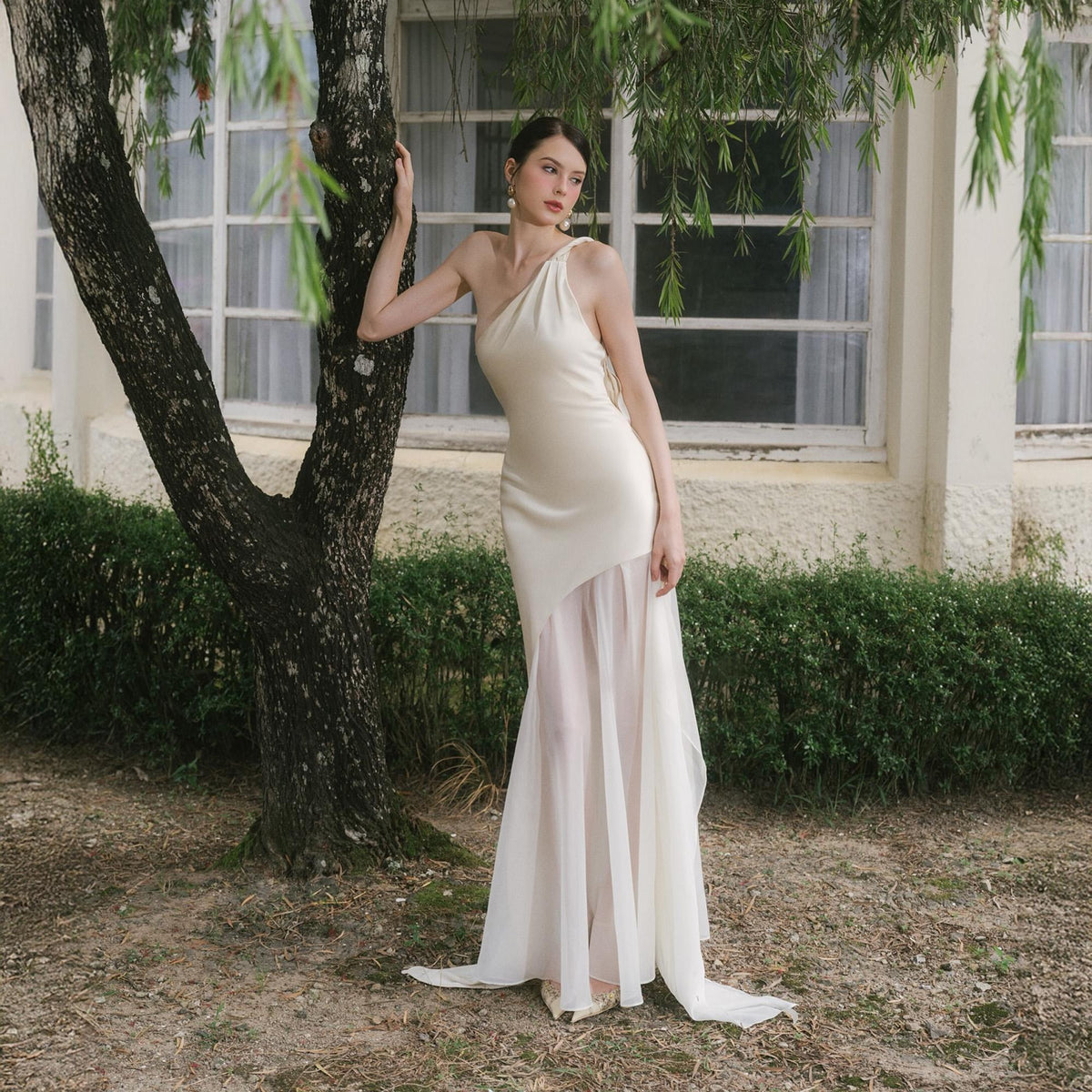 May include: An ivory-colored, one-shoulder gown with a flowing, sheer skirt. The dress has a fitted bodice and a long, asymmetrical skirt. The model is standing near a tree in front of a building with windows. The dress is suitable for a wedding or formal event.