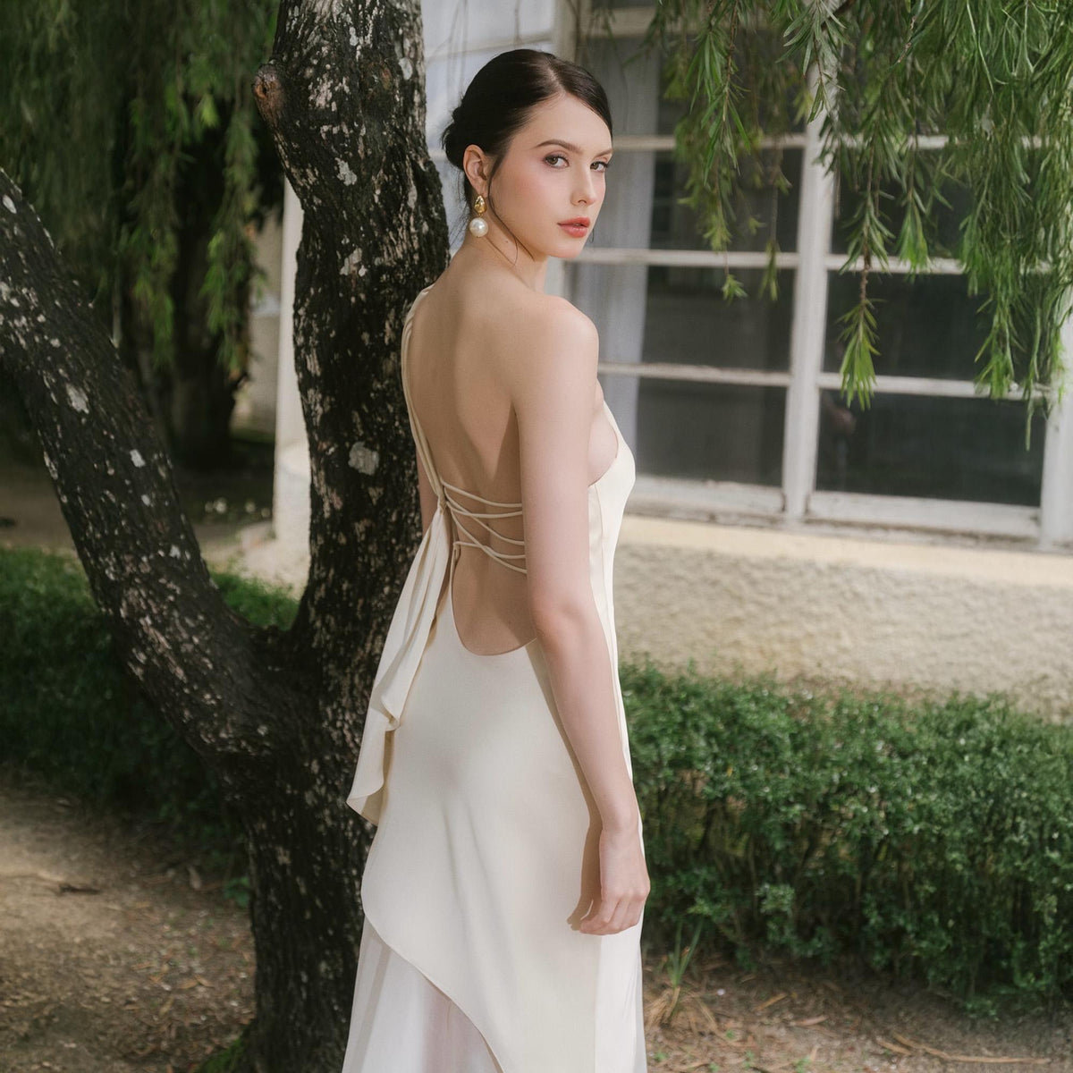 May include: A cream-colored, backless dress with spaghetti straps and a flowing skirt. The dress features a low back with criss-cross straps and a draped fabric detail. The model is wearing pearl earrings and looking over their shoulder.