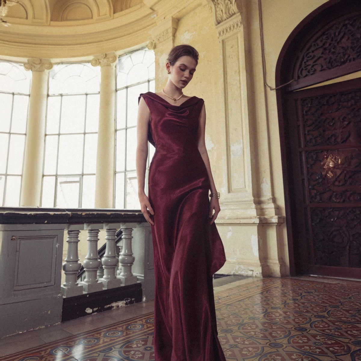 May include: A burgundy-colored, floor-length gown with a cowl neckline. The dress has a flowing, silky appearance and is worn in a grand, ornate building with large windows and decorative architectural details. The dress is a formal wear item.