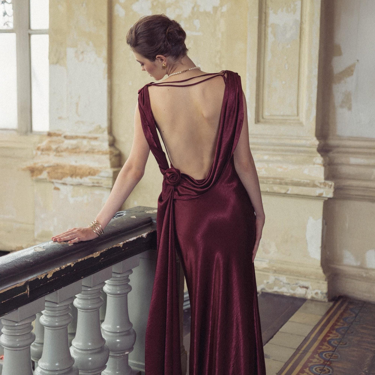 May include: A burgundy satin evening gown with a low draped back and a rose detail at the waist. The dress has a flowing skirt and thin straps. The model is standing near a staircase with a white railing.