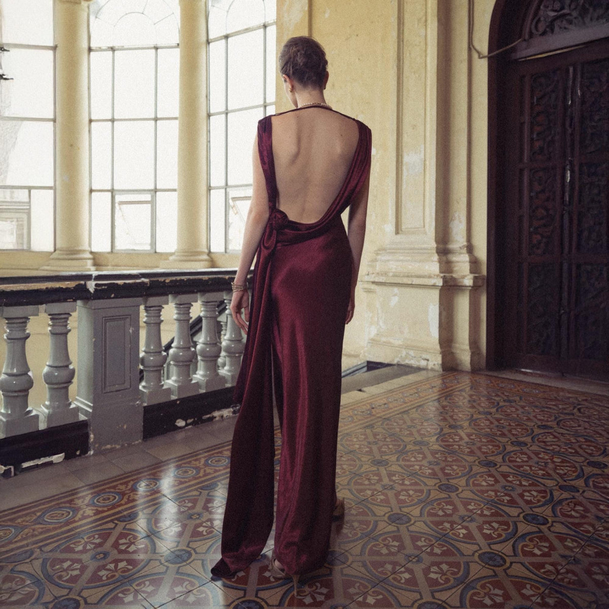 May include: A burgundy, floor-length gown with a low back and draped fabric. The dress features a flowing train and is worn in a well-lit, ornate hallway with a patterned tile floor.