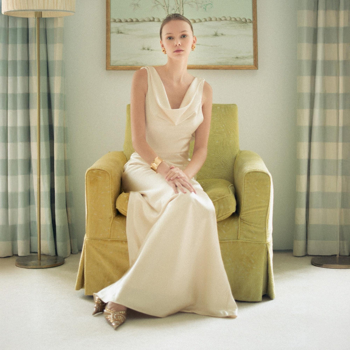 May include: A floor-length cream-colored sleeveless gown featuring a draped cowl neckline and a fluid, floor-sweeping skirt. The garment is paired with gold-toned earrings and a wide gold-toned cuff bracelet, complemented by pointed-toe gold shoes.