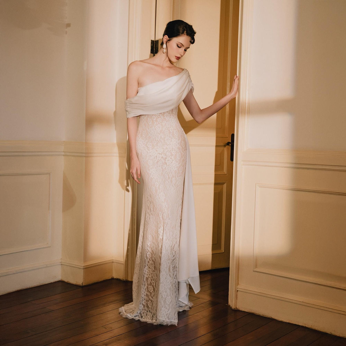 May include: A floor-length, off-the-shoulder gown featuring a white, draped fabric bodice and a fitted, floor-length skirt made of intricate white lace. The design includes a single shoulder strap and a long, flowing fabric panel extending from the side.