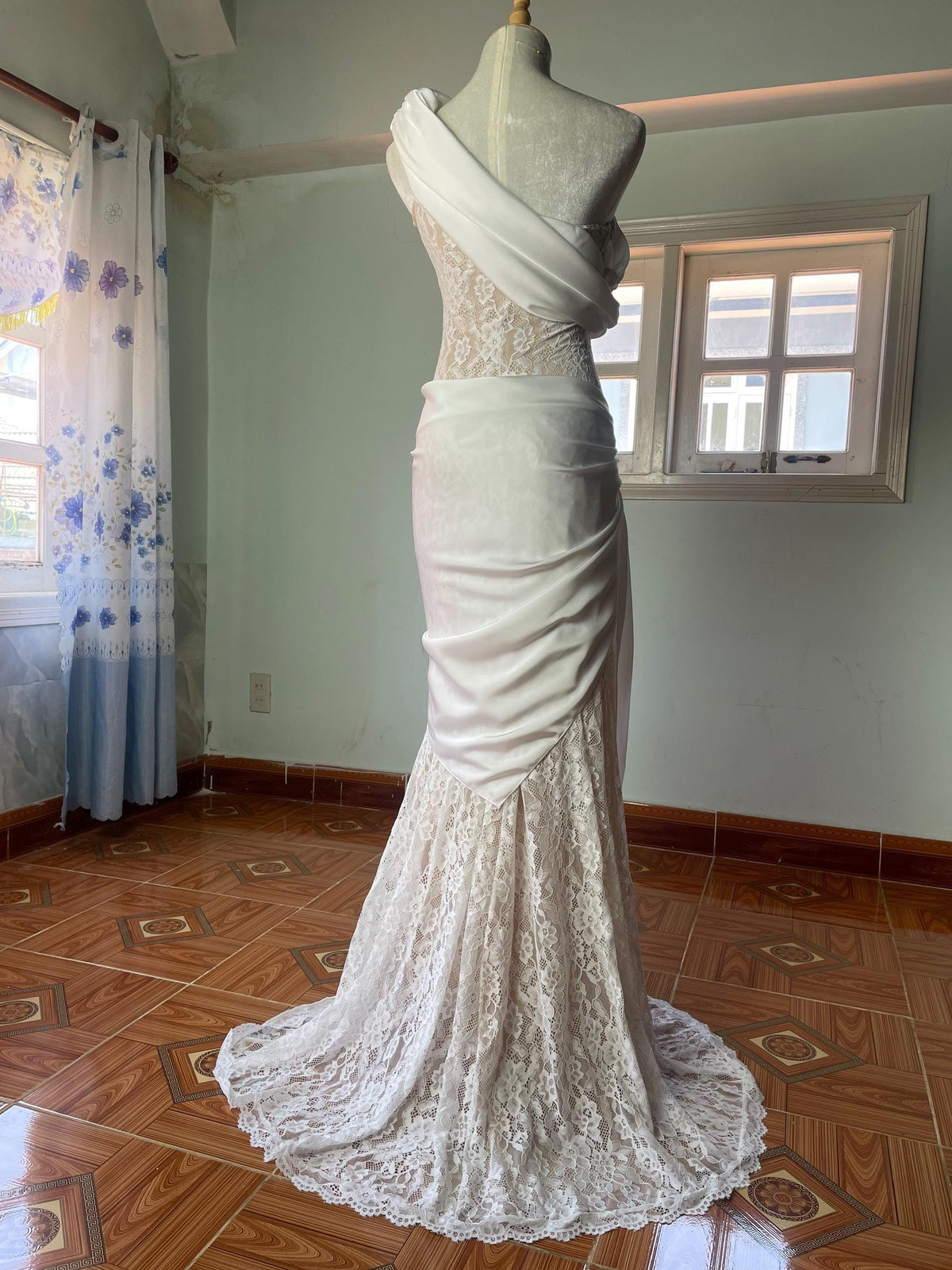 May include: A floor-length white gown featuring a one-shoulder neckline with draped fabric across the bodice. The garment combines a solid white fabric overlay with an intricate white lace underlay that extends to a full-length skirt and hem.