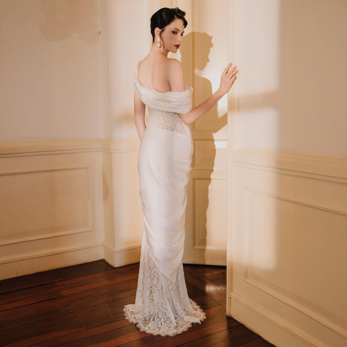 May include: A floor-length white gown featuring an off-the-shoulder neckline with draped fabric across the bodice. The garment includes a fitted waist section and a skirt that transitions into a detailed white lace hemline at the bottom.