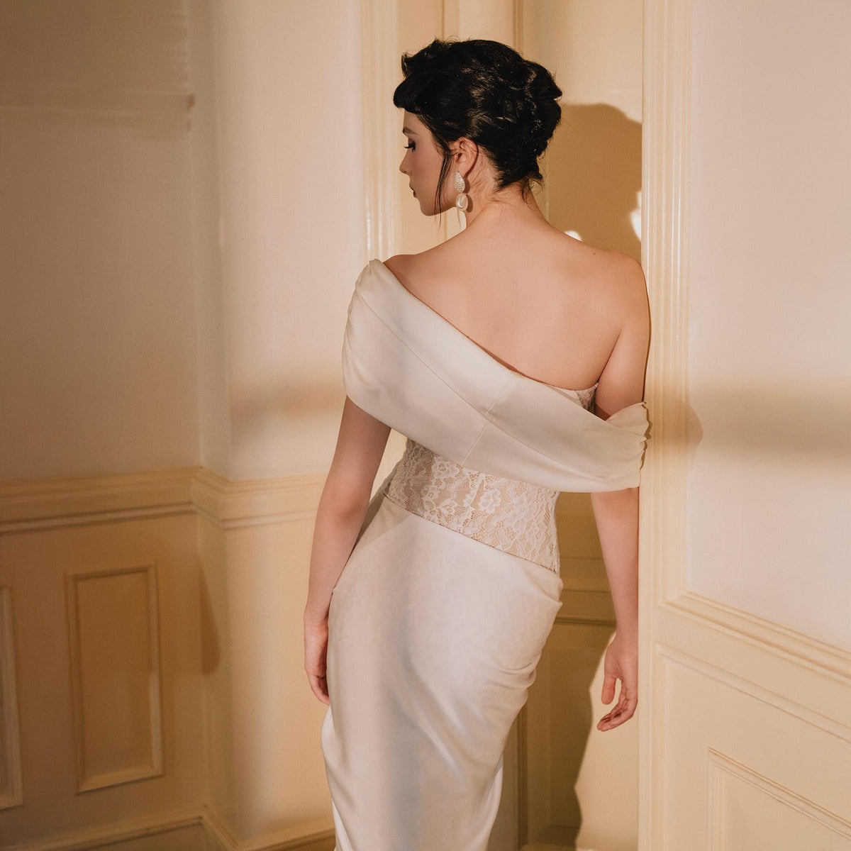 May include: A floor-length off-the-shoulder ivory gown featuring a smooth satin bodice and a sheer lace midsection. The dress has a draped neckline that wraps around the upper back and shoulders, transitioning into a sleek, solid satin skirt.