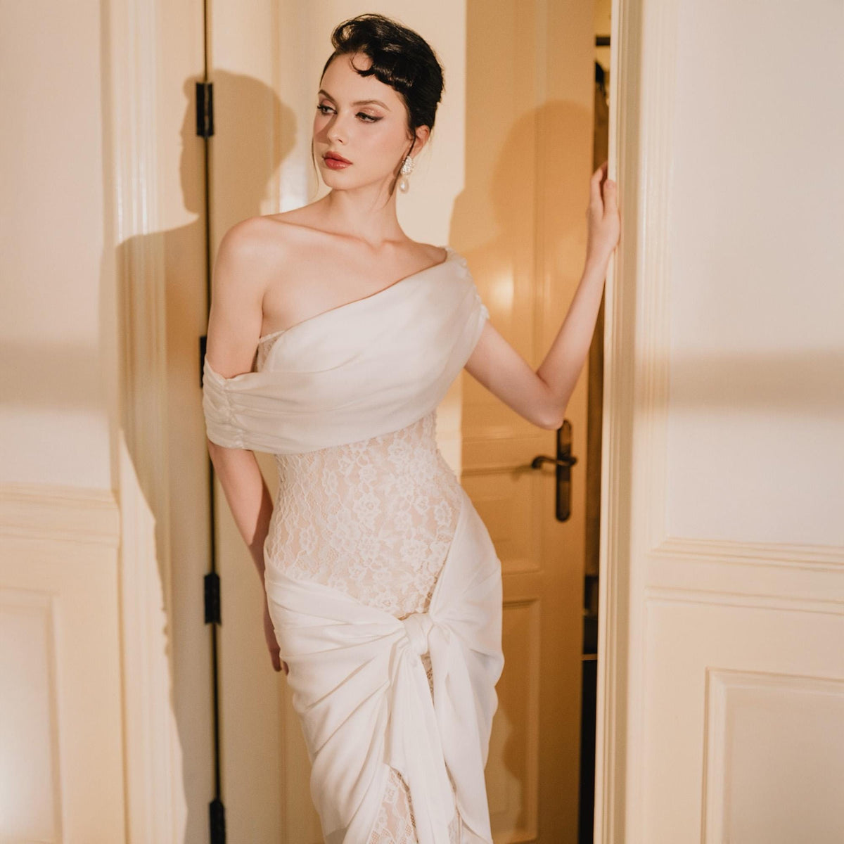 May include: A floor-length white dress featuring a one-shoulder draped bodice, a sheer lace overlay on the torso, and a gathered fabric detail tied at the front waist. The garment has a fitted silhouette and a smooth, flowing skirt section.
