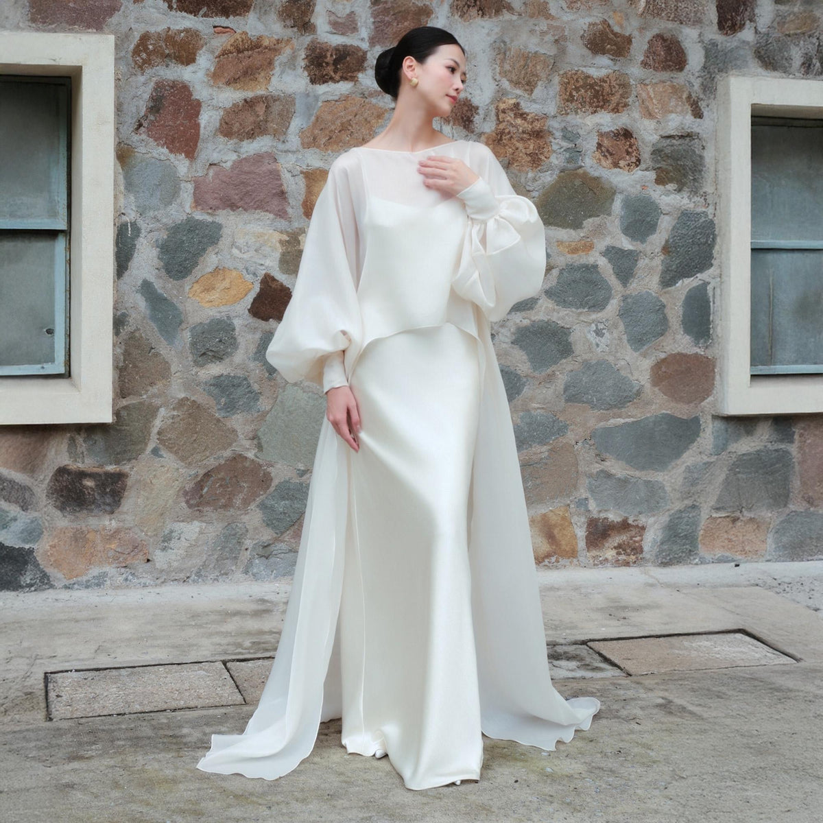 May include: An ivory-colored bridal gown with a flowing skirt and a sheer, long-sleeved top. The top features voluminous sleeves and a boat neckline. The dress is made of a silky material and has a long, flowing train. The background is a stone wall.