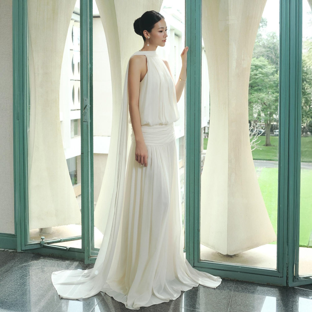 May include: A floor-length, ivory-colored dress with a flowing skirt and a high neckline. The dress features a gathered waist and a train. The woman is standing near a window with sheer curtains. The dress is made of a lightweight fabric.