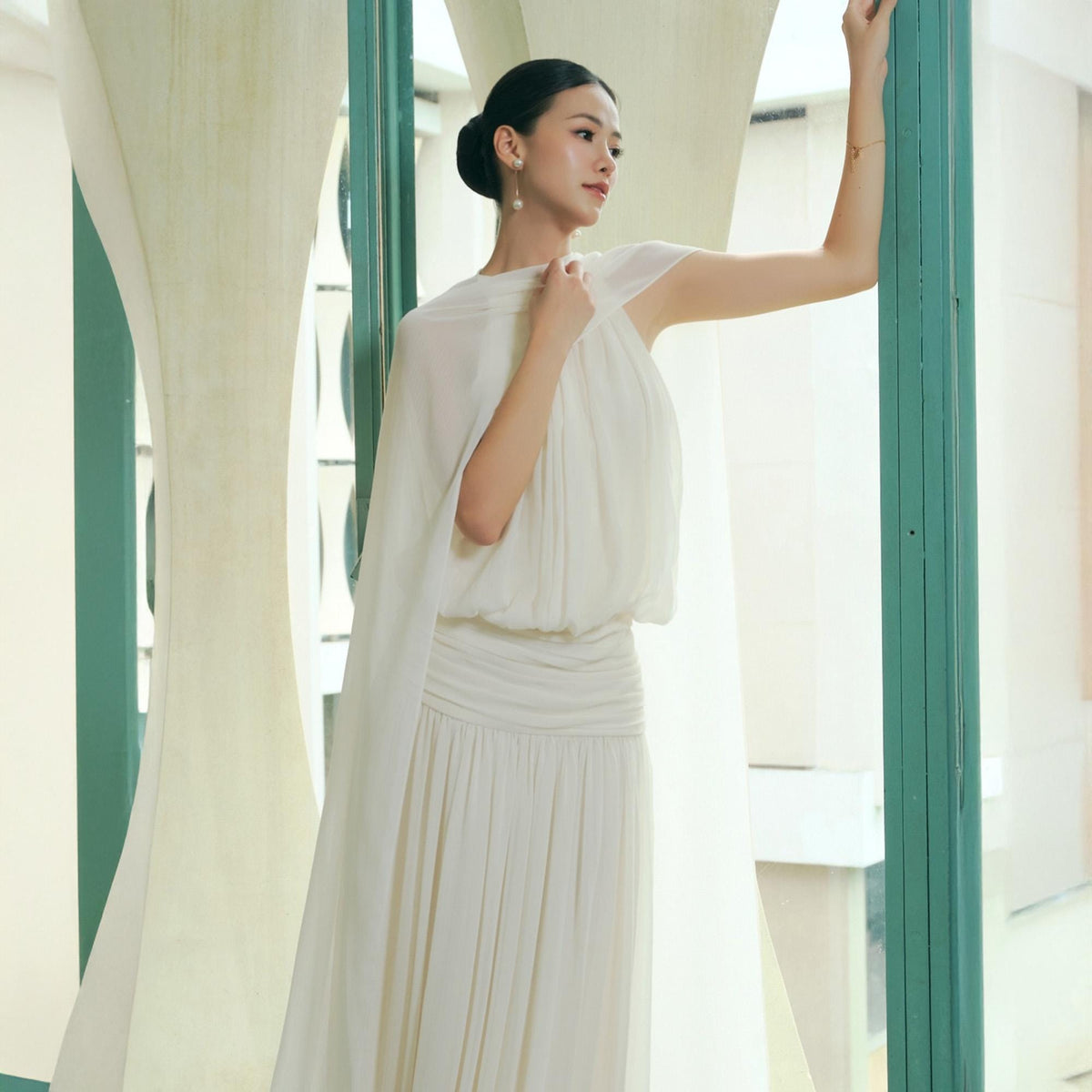 May include: A woman in a white dress with a draped shoulder detail and pleated skirt. The dress has a flowing, elegant design. The woman is standing near a window with a green frame and a light-colored, patterned background.